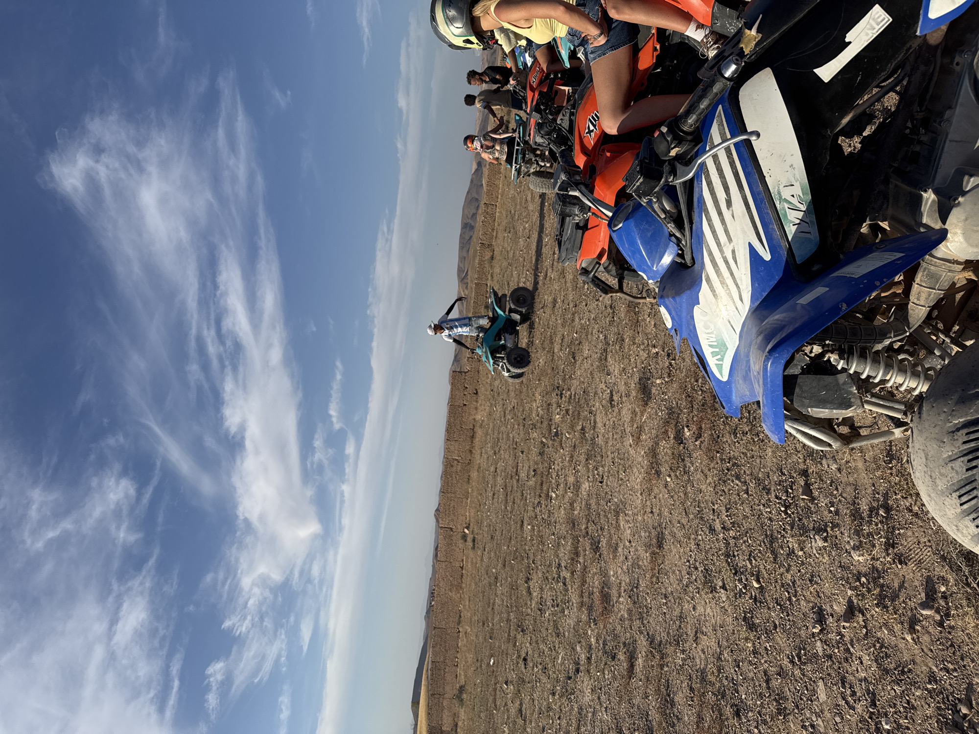 In Lalla Takarkoust, Morocco, a group of individuals are gathered around several all-terrain vehicles (ATVs) in a dry, open field under a bright blue sky with wispy clouds. In the foreground, a blue and black ATV is partially visible, with a person's legs and feet in denim shorts and white socks wearing sneakers seated on an orange ATV behind it. Other ATVs, including an orange one and a teal one, are parked nearby.
A man in a white and blue striped shirt and white cap stands on the teal ATV, gesturing with his arms. Several other individuals, some wearing helmets, are either seated on or standing near the ATVs. In the background, a low, rough-textured wall made of earth or stone runs horizontally across the landscape. Beyond the wall, rolling hills and mountains are visible under the clear sky. The ground is dry and covered with pebbles and sparse vegetation. The overall scene suggests an outdoor recreational activity, likely a tour or adventure involving ATVs in a rural Moroccan setting.