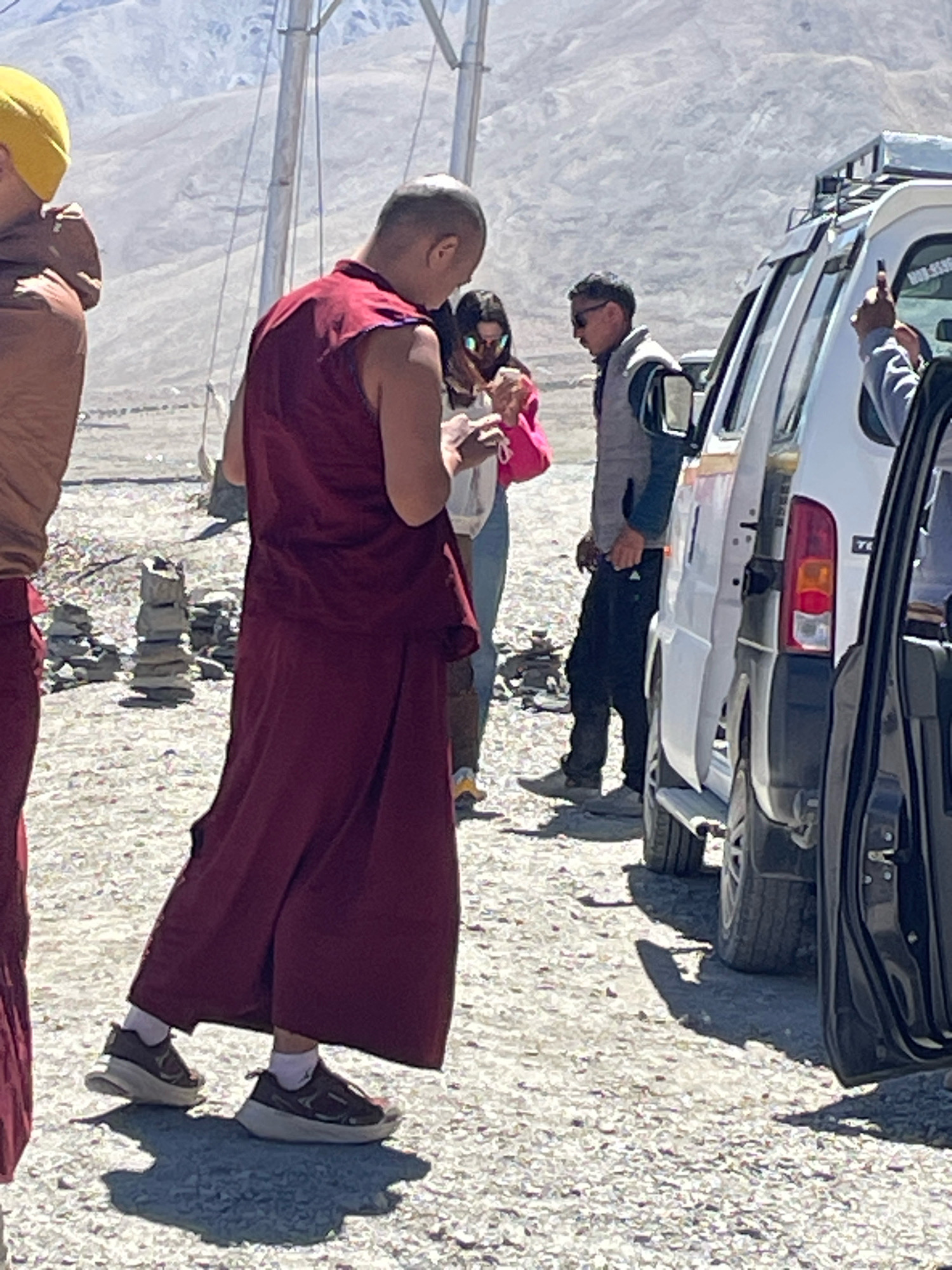 In an outdoor, mountainous environment in the city of Unknown, India, a Buddhist monk wearing a maroon robe and yellow cap stands next to another individual dressed in similar attire. A third person, a woman in casual clothing and sunglasses, holds a pink bag and appears to be interacting with the monk. To the right, a white SUV with a roof rack is parked, and a black car is partially visible on the far right. The ground is covered in gravel, and stacks of stones are present in the mid-ground. The scene is illuminated by bright sunlight, casting distinct shadows.