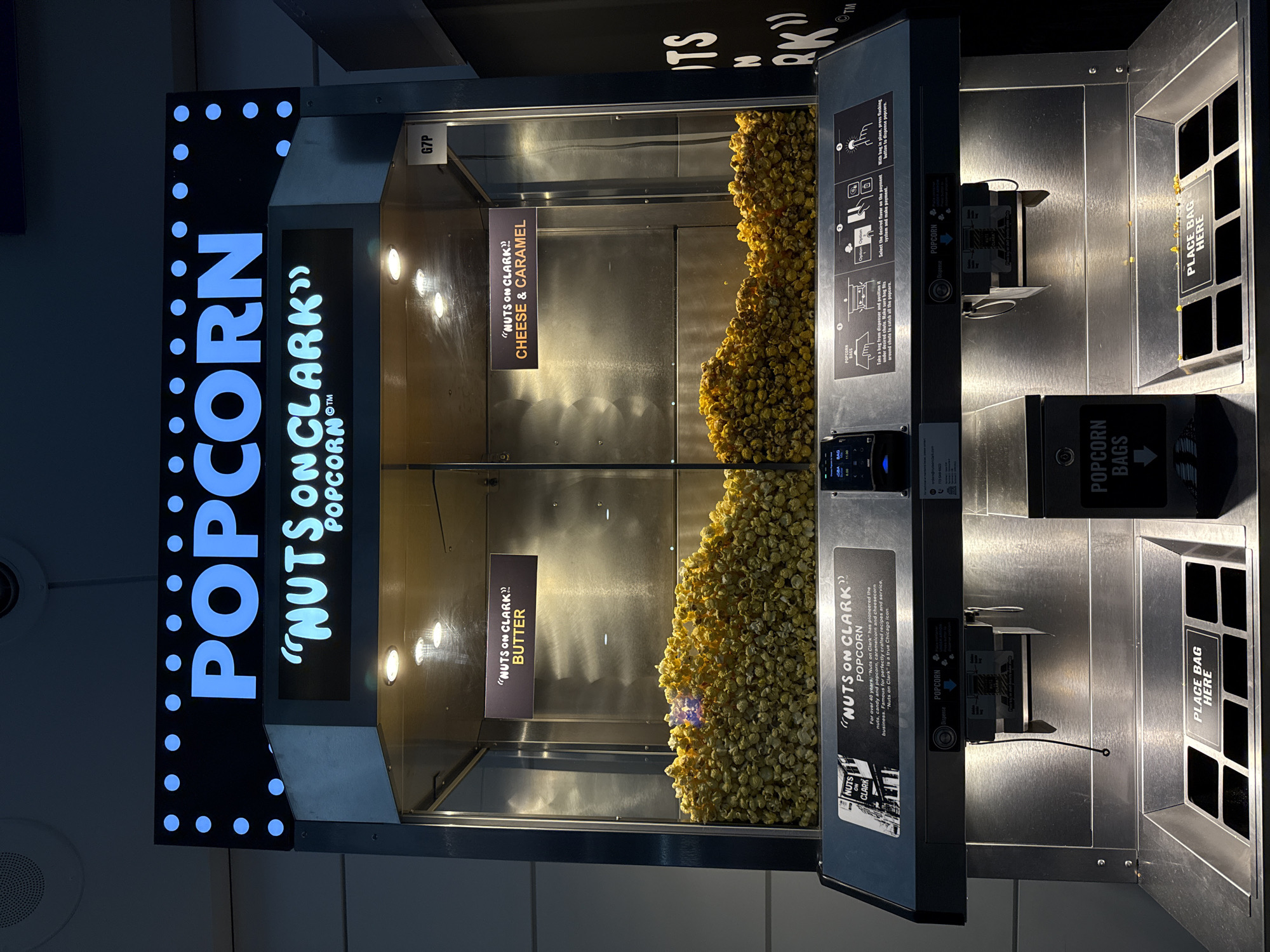 This image shows a "Nuts on Clark" popcorn vending machine. The vending machine has two different flavors of popcorn available: butter and cheese & caramel. The location of the vending machine is not explicitly indicated. However, the brand is "Nuts on Clark", a famous Chicago popcorn brand. The machine is fully automatic. Instructions on the machine explain that you can take a bag from the dispenser, select a flavor and make a payment, and press the flashing button to dispense the popcorn.