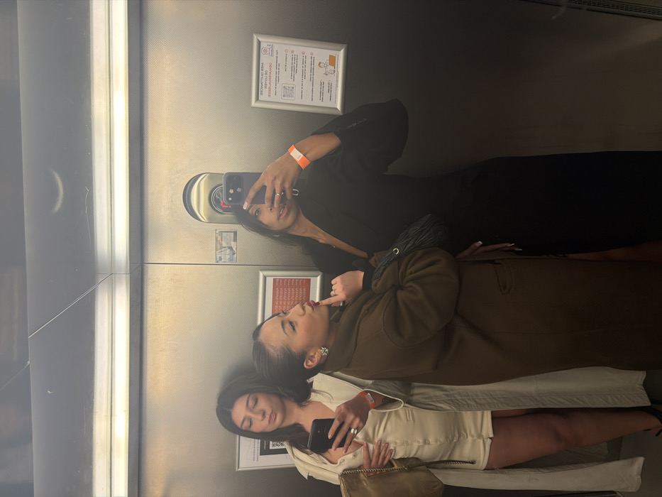 Three women are in an elevator taking a selfie in the mirror. One woman wears a black blazer and holds a phone up to take the picture. Another has a brown coat and her finger near her lips. The third wears a tan dress and holds a phone. All three have wristbands. There are elevator buttons and safety posters visible on the mirror.