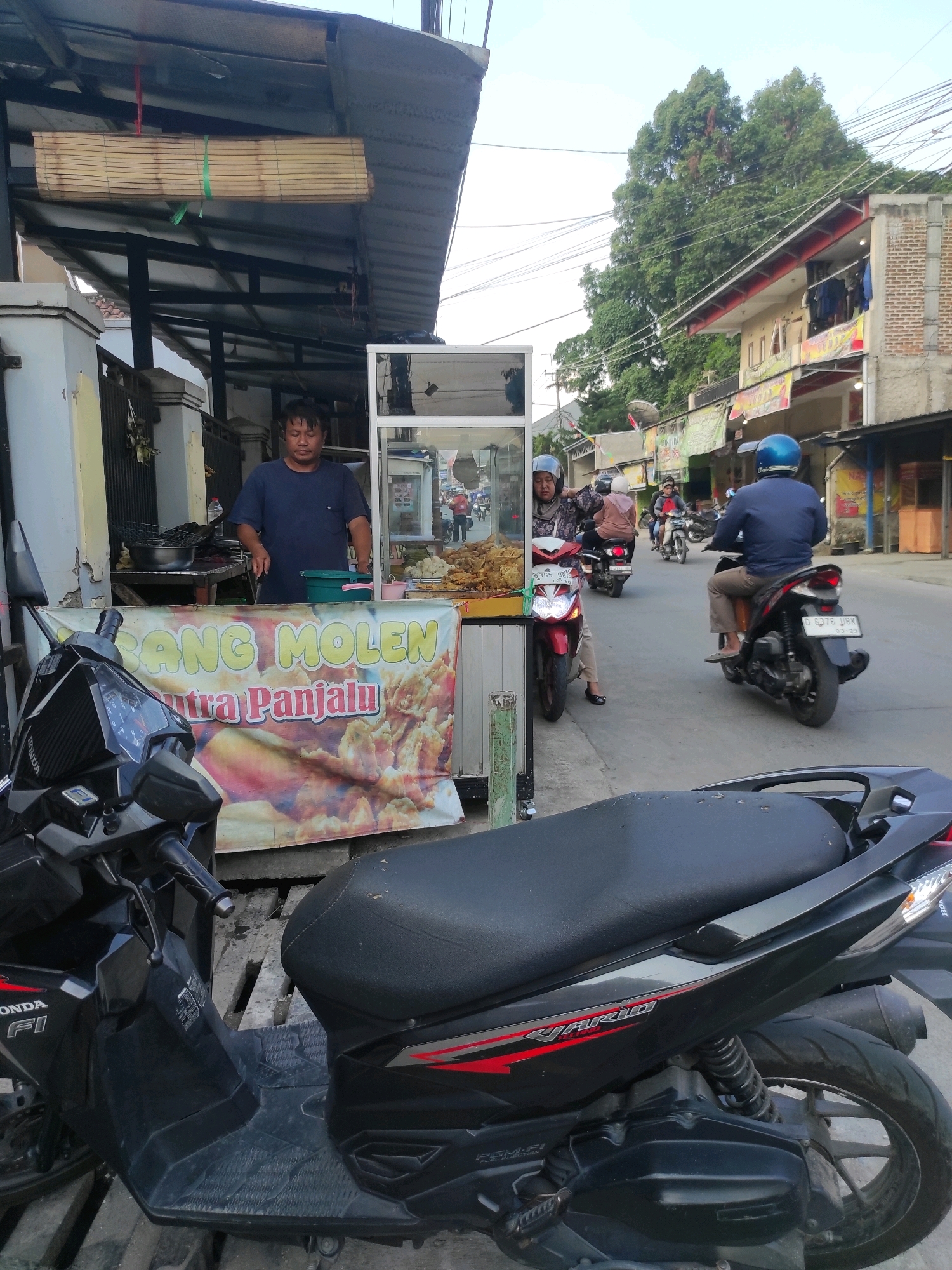 This is a street-level, medium-distance shot of a food stall in Padalarang, Indonesia. The stall appears to be a small, open-air setup with a metal roof and a glass display case. A man in a dark blue shirt is standing behind the counter, holding a knife and a green bucket. In front of the stall, a large banner displays the text "SANG MOLEN" and "Sutra Panjalu," with an image of fried food.
In the foreground, the back half of a black Honda Vario scooter is visible, parked on a wooden pallet. The scooter has "HONDA FI" written on its side.
On the street behind the stall, several people on motorbikes are passing by. One woman in a headscarf is on a red scooter. Further down the street, a man in a blue helmet is riding a black scooter. Other motorbikes and people are present in the background, along with buildings and trees. The sky is overcast. The overall scene depicts a typical street food vendor in an urban Indonesian setting.