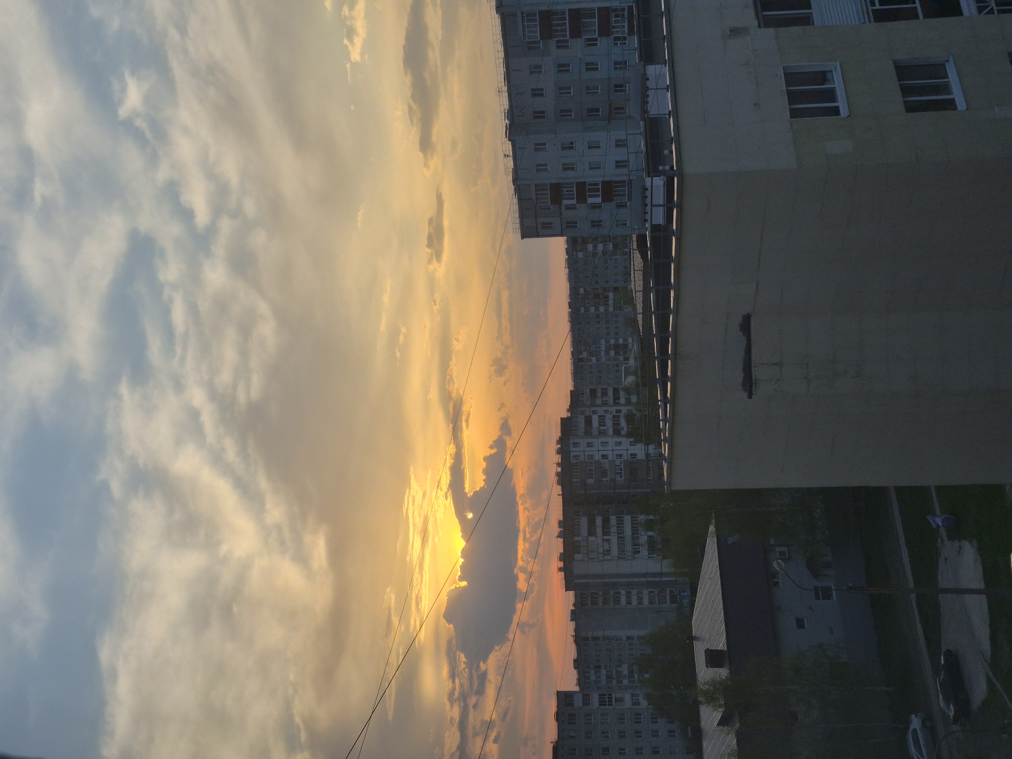 The image depicts a sunset over a city in Unknown, Unknown. The sky is filled with clouds ranging in color from white to orange. Buildings, mostly apartment complexes, can be seen silhouetted against the sky. A few power lines are visible in the sky.