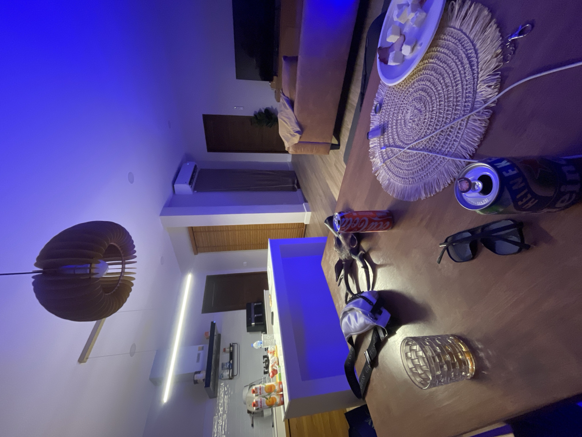 The image depicts a dimly lit interior scene, possibly a residential living or dining area, with strong blue ambient lighting. In the foreground, a dark wooden table is laden with several objects. These include a glass containing an amber liquid, a pair of black sunglasses, a white fanny pack with black straps, a can of Coca-Cola, and an open can of Heineken beer. A woven placemat sits on the table, with a white plate holding several cubes of cheese and some sliced meat. A charging cable extends from the placemat towards the right side of the frame.
In the midground, a modern kitchen island with a white countertop is illuminated with the same blue light, and several bottles of juice are visible on top. Above the island, a sleek range hood is mounted on a tiled backsplash. Further back, a brown leather sofa is positioned in front of a large black television screen. A set of wooden blinds is visible on a door or window to the right. An air conditioning unit is mounted high on the wall in the background. The overall impression is that of a relaxed, possibly late-evening setting. The location specified as Baan Plai Laem, Thailand, provides a geographical context, suggesting the scene is within this area.
