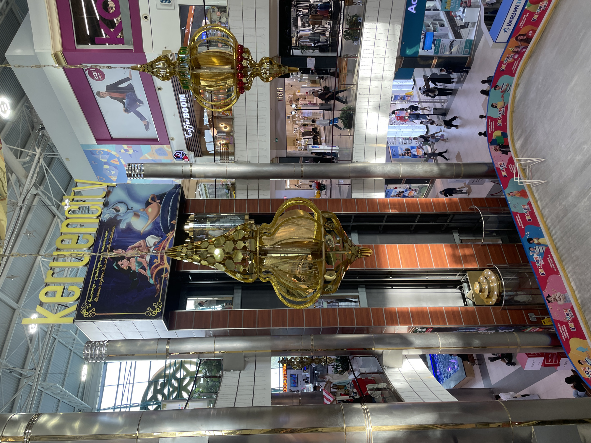 This is an indoor shot of a multi-level shopping mall, likely in Aktobe, Kazakhstan. The scene is dominated by large, metallic columns and a central glass elevator shaft. Overhead, large yellow lettering spells out "Keruen City." Several large, ornate golden lamps hang from the ceiling, suggesting festive or decorative lighting.
On the upper levels, shop fronts are visible, including "Kari" and "Coffee BOOM." People are seen browsing or walking through the mall on various levels. In the foreground, a section of an ice rink is visible, bordered by a colorful banner with the "Keruen Park" logo and images of families and children. The overall impression is a brightly lit, modern commercial space designed for shopping and leisure activities.