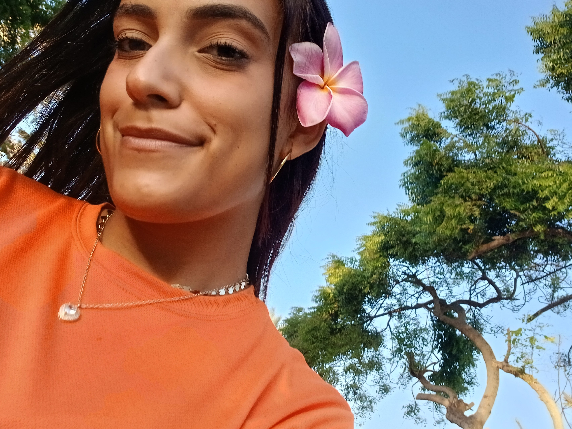 A young woman with dark hair and light skin smiles at the camera. A pink frangipani flower is tucked behind her left ear. She wears a gold hoop earring, a gold coin necklace, and a gold necklace with a pendant. She is wearing an orange t-shirt. Behind her are trees with green leaves against a blue sky. The image appears to have been taken outdoors during daylight hours. The location is possibly Playa El Agua, Venezuela.
