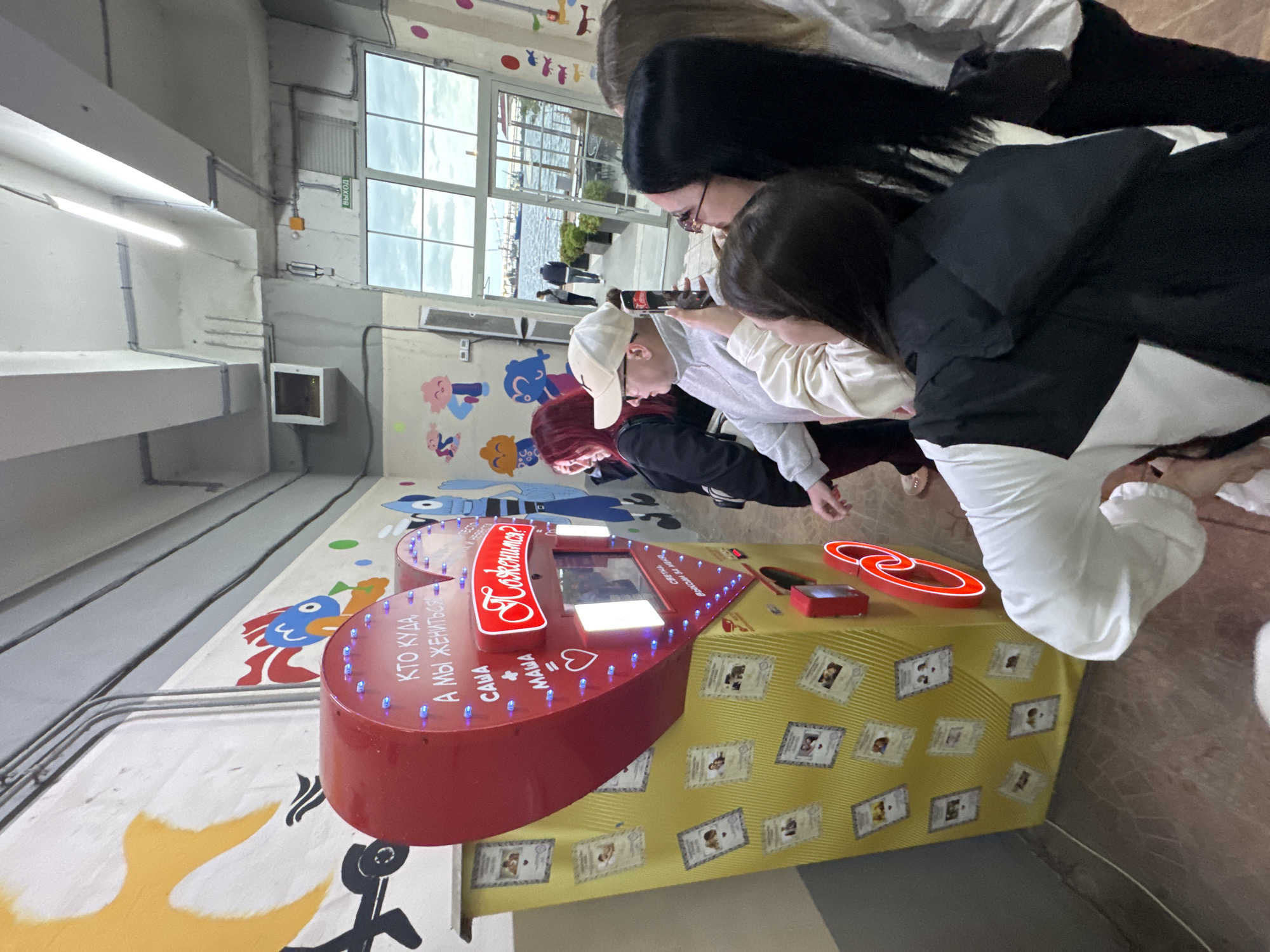 In Saint Petersburg, Russia, a group of young adults is gathered around a large, heart-shaped photo booth decorated with lights and images of couples. The booth, painted red and yellow, is adorned with numerous small photos of smiling couples, suggesting it's a romantic attraction. Text on the booth reads "Кто куда, а мы жениться!" which translates to "Where everyone goes, we're getting married!". The environment appears to be an indoor public space, possibly a pier or promenade, with large windows in the background revealing a cloudy sky and a body of water with boats. The people, dressed casually, are interacting with the photo booth, with one person taking a photo of the booth with their phone. The overall atmosphere suggests a lighthearted and celebratory occasion.