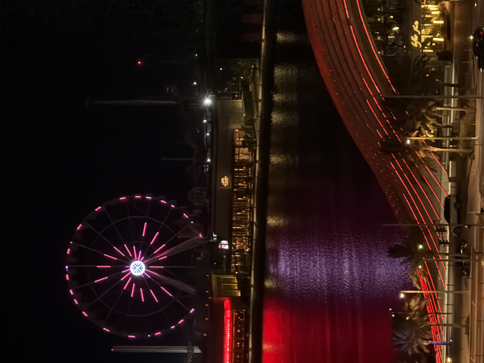 The image captures a nighttime view of a waterfront area in an unknown city. A large Ferris wheel, illuminated with pink and white lights, dominates the upper left portion of the frame. Below the Ferris wheel, a row of buildings lines the waterfront, featuring lit windows and glowing red signage on the left. The water reflects the vibrant pink and purple lights from the Ferris wheel and buildings. In the foreground, a futuristic-looking bridge or elevated walkway, adorned with numerous red lights, curves across the scene. Palm trees are visible along the walkway, and several cars are parked or driving on a road below. The overall atmosphere is one of urban nightlife and illuminated architecture.