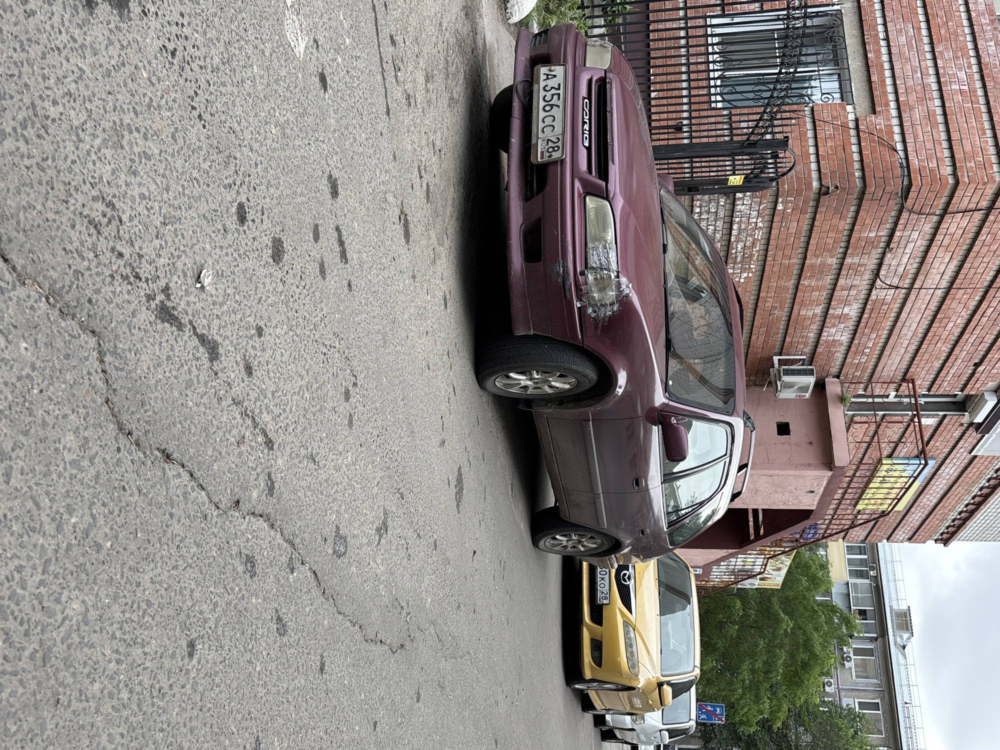 This is a photograph of a street scene in Blagoveshchensk, Russia. The image is taken from a low angle, looking upwards at several parked cars. The dominant subjects are vehicles, including a maroon car with a visible license plate reading "A 356 CC 28 RUS" and the word "CARIB" on its grille. Behind it is a yellow car with a license plate that appears to read "60 KO 28". Further cars are visible in the background, though less distinct.
The setting is an urban environment, characterized by a cracked asphalt road and a red brick building on the right side of the frame. The building has several windows, some covered with black metal bars, and air conditioning units are visible. There are also metal railings and a sign with colorful graphics on the building's facade. Lush green trees are present behind the cars, suggesting a potentially warm season.
The time of day appears to be daytime, and the weather seems overcast, as indicated by the diffused light and the overall muted tones of the sky. No people are visible in the frame. The scene depicts a quiet street with cars parked along the side. There are no overt signs of activity or emotion, conveying a sense of stillness.
