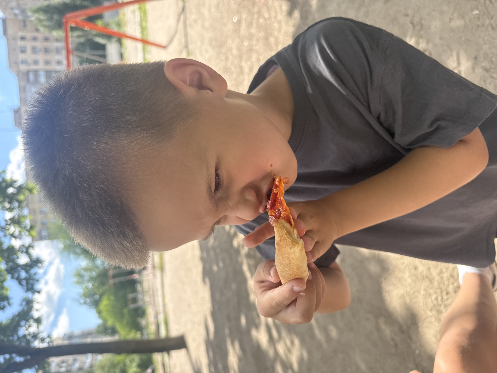 A young boy, estimated to be between 4 and 6 years old, is shown in a close-up profile shot, focused on his face as he eats a piece of pizza. He has short brown hair and is wearing a dark gray t-shirt. His eyes are narrowed in concentration as he brings the pizza to his mouth, and there are traces of red sauce on his lips and chin. His left hand is holding the pizza crust, while his right hand is near his mouth.
The background suggests an outdoor urban environment, likely a playground or park area. In the mid-ground, a bright orange swing set is visible, and further back, apartment buildings can be seen. The ground appears to be a gravel or dirt surface, with shadows from trees indicating it is daytime. The lighting suggests a sunny day.
The overall scene conveys a candid moment of a child enjoying a snack. There are no other individuals clearly identifiable in the frame, although a portion of another person's arm is visible in the lower left corner, suggesting they are holding the pizza for the boy or are with him. The context implies this is a casual, everyday scene.
