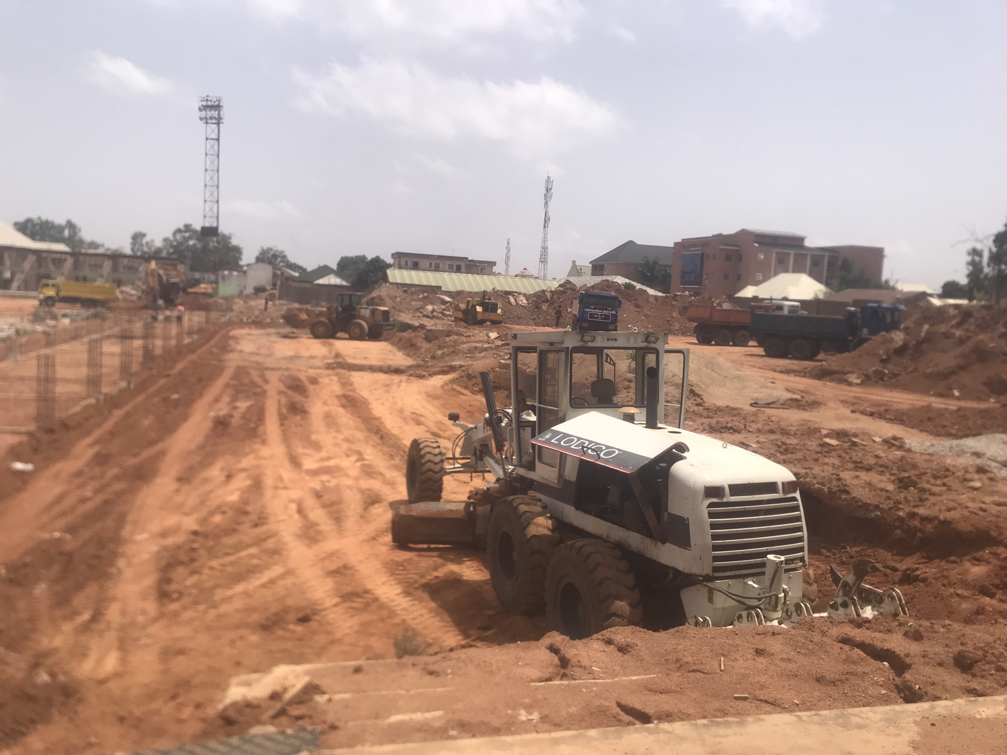 The image depicts a construction site in Jos, Nigeria. The foreground features a white grader, partially submerged in red soil. In the background, other construction vehicles, including bulldozers and dump trucks, are visible along with ongoing work. Reinforcement bars suggest ongoing construction. A tall communications tower stands in the distance. The environment is dusty and the sky is overcast. Buildings are scattered in the background.