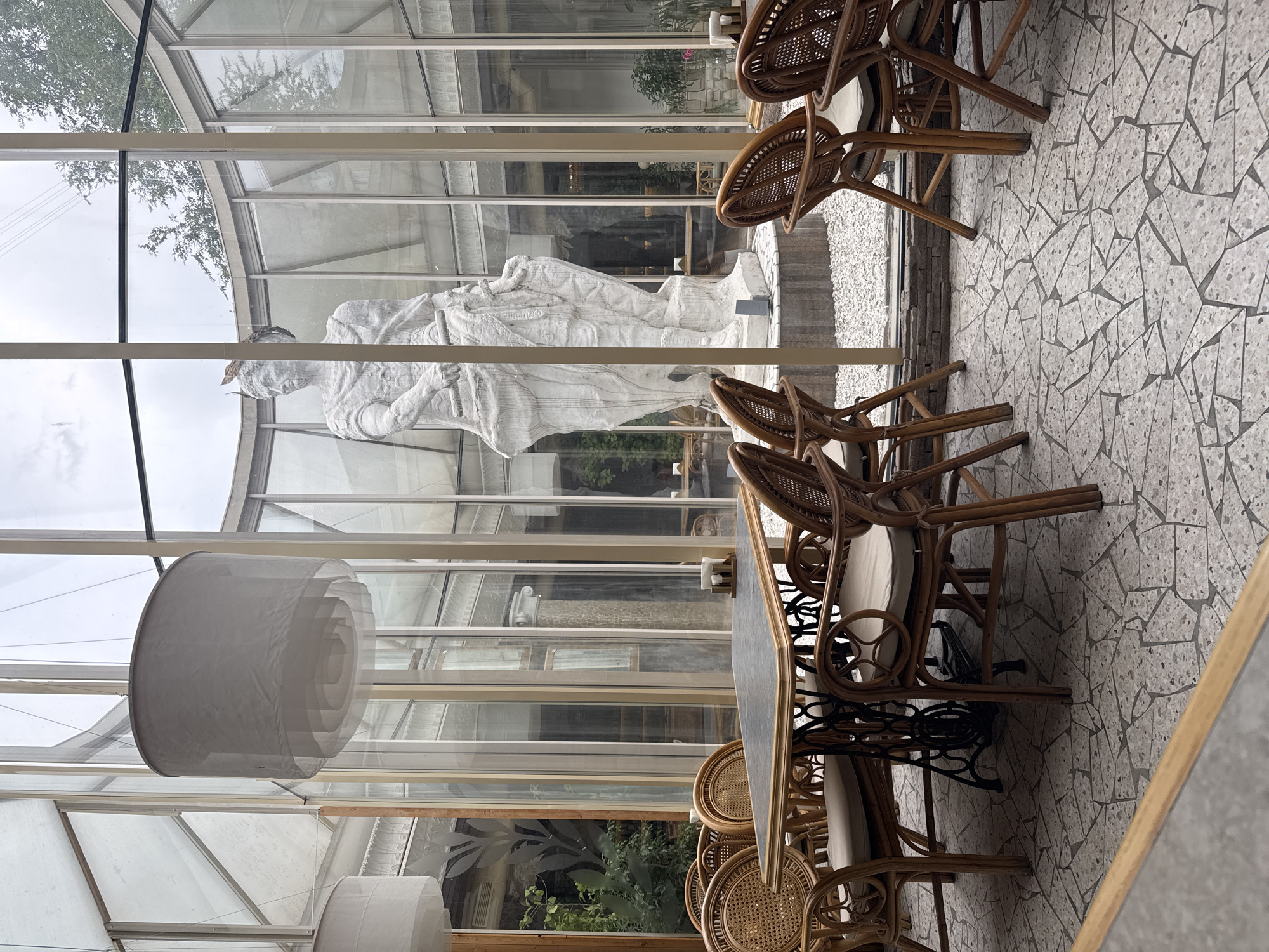 This image depicts the interior of a glass-enclosed space, possibly a restaurant or atrium, with a large, white classical statue as a central focal point. The floor is covered in a mosaic of light-colored tiles with darker grout. Several tables with rattan chairs surround the central area. One table in the foreground features a dark, rectangular top and a black, ornate base resembling a sewing machine. The chairs are also made of rattan with light-colored cushions. In the background, through the glass walls and ceiling, trees and an overcast sky are visible. The statue appears to be of a robed figure holding a staff or pole, with some sort of headpiece. There are no people visible in the image. The lighting is diffused, suggesting an overcast day or an indoor environment with artificial lighting.