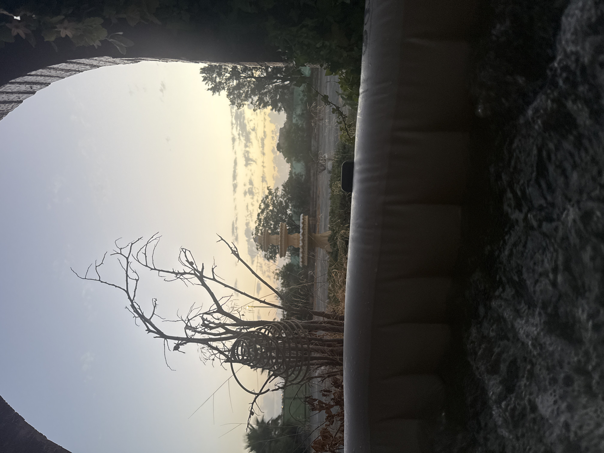 The image captures a serene outdoor scene, viewed from within a hot tub. The foreground is dominated by the bubbling water of the hot tub, creating a textured, dark surface. Beyond the tub, a barren, thorny branch structure stands prominently, silhouetted against a soft, dusky sky. In the mid-ground, a decorative tiered fountain is visible, surrounded by dry vegetation and a scattering of trees. The background reveals more trees and a fence line, suggesting an open, perhaps rural, environment. The scene is framed by a dark, arched structure on the right, with ivy creeping down its side, and a similar, but less defined, arch on the left. The lighting indicates either dawn or dusk, with warm, pale tones illuminating the sky and casting soft shadows. The location is identified as Corvera, Spain. There are no people or active interactions depicted in the scene.