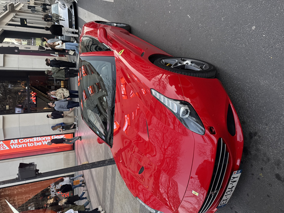 A bright red Ferrari convertible, license plate EX-134-VY (French registration), is prominently positioned in the foreground on an asphalt road. Its black retractable hardtop is closed, and reflections of bare tree branches are visible on its glossy surface. The vehicle appears stationary, angled slightly towards the right. In the mid-ground, a second Ferrari, silver or light gray and also a convertible, is stationary further down the street, with a partially visible license plate EX-681-TE.
To the left, a wide paved sidewalk borders a multi-story building housing a Nike store, identifiable by its logo and a vertical red banner displaying "All Conditions Gear." The building features modern glass and light-colored facade elements. Numerous casually dressed pedestrians are present on the sidewalk, walking in various directions or standing in groups. A dark traffic signal pole stands on the left side of the road, near the sidewalk. The architectural style of the buildings and the presence of a flagship Nike store confirm the location as Avenue des Champs-Élysées in Paris, France. The scene is illuminated by natural daylight.