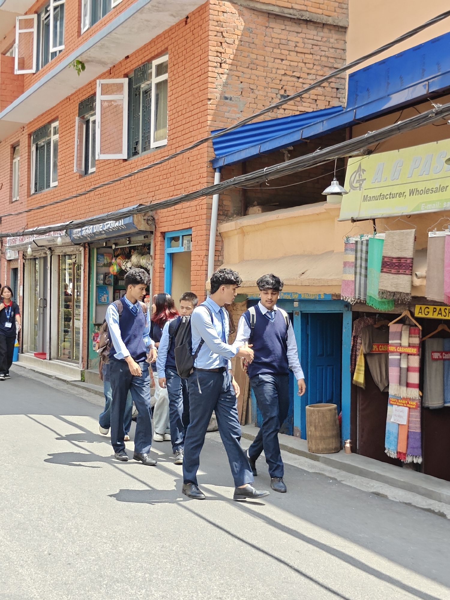 A group of young males, dressed in school uniforms consisting of light blue collared shirts, dark blue V-neck sweaters, and dark blue trousers, are walking along a paved street. The group appears to be engaged in conversation. One individual in the foreground, facing right, has his left hand extended, as if in mid-gesture. They are walking past shops with open storefronts displaying various items. One shop has colorful textiles hanging outside, including scarves and shawls, with signage indicating "100% BABY YAK" and "Hand Made in Nepal." Another shop features what appear to be toys or decorative items behind glass. The buildings are constructed of brick and have multiple windows. Power lines are visible above the street. The scene is bathed in sunlight, casting distinct shadows on the pavement. The location appears to be a street within Kathmandu Metropolitan City, Nepal, based on the architectural style and signage.