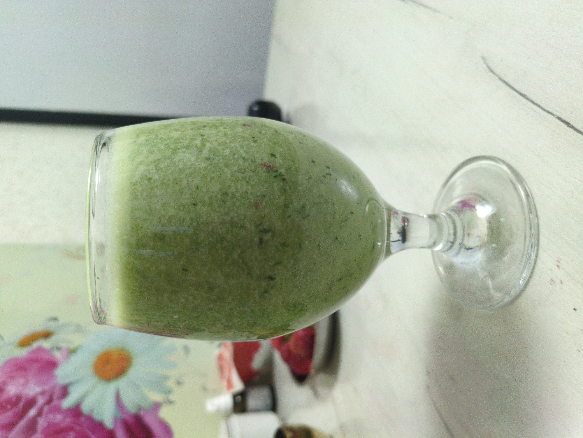 A close-up, eye-level, medium shot shows a clear glass goblet filled with a thick, pale green smoothie, positioned on a light-colored, textured surface. The smoothie is opaque and appears to contain small flecks of darker green material, possibly herbs or fruit pulp. The goblet has a stem and a circular base. In the blurred background, a portion of a fabric with a floral pattern, featuring white daisies with orange centers and pink flowers, is visible on the left. To the right of the smoothie, a small, dark, rounded object is out of focus. Further to the left and in the background, other out-of-focus objects include what appear to be bottles and a red bowl containing red fruit, possibly strawberries. The overall lighting suggests an indoor setting, likely a kitchen or dining area. The image does not contain any information or visual cues that would suggest a specific location in Kulebaki, Russia.