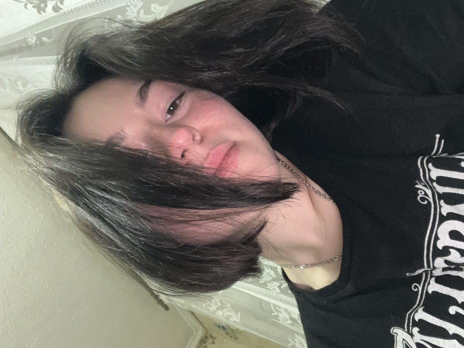 A young person with dark hair and fair skin lies in repose. Strands of hair fall across their face, partially obscuring one eye. The person wears a black t-shirt with stylized white print. A delicate silver chain rests around their neck, and a patterned wallpaper is visible in the background. The image is composed at an angle, giving a sense of informality and relaxation.