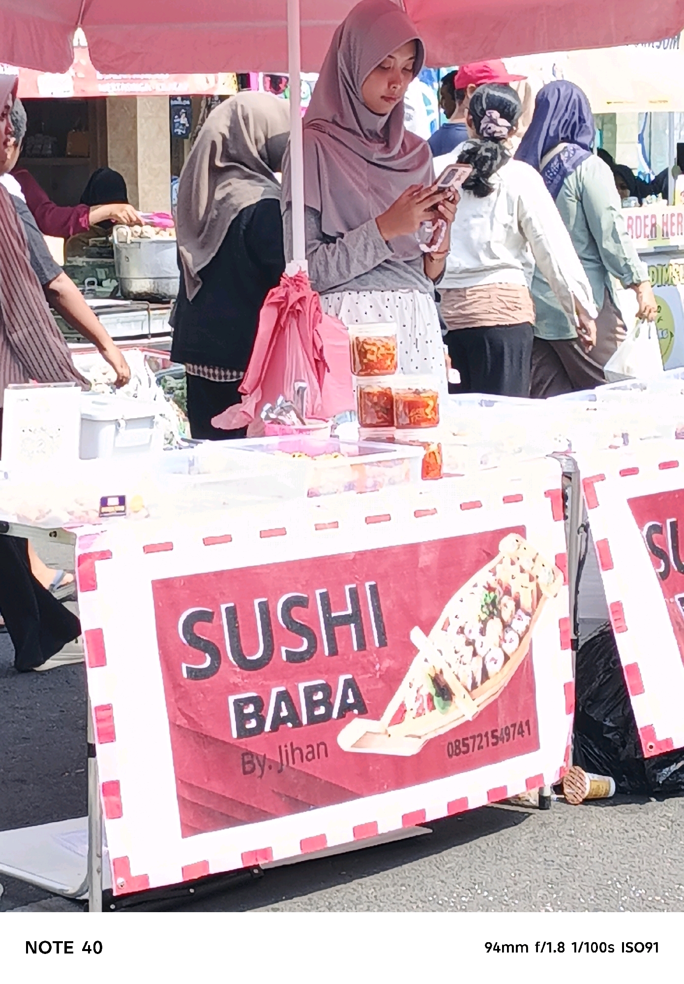 A street vendor is selling sushi from a stall under a pink canopy. The stall features a prominent red and white sign with the text "SUSHI BABA By. Jihan" and a phone number. Several containers of what appear to be sushi or related food items are displayed on the stall. A young woman wearing a grey hijab and a patterned skirt is visible at the front of the stall, looking at her phone. Other individuals, including women wearing hijabs and men, are present in the background, some engaged in transactions or preparing food. The environment suggests a bustling outdoor market or street scene in Cilacap, Indonesia, with a paved ground surface and other stalls or shops visible behind the main subject.