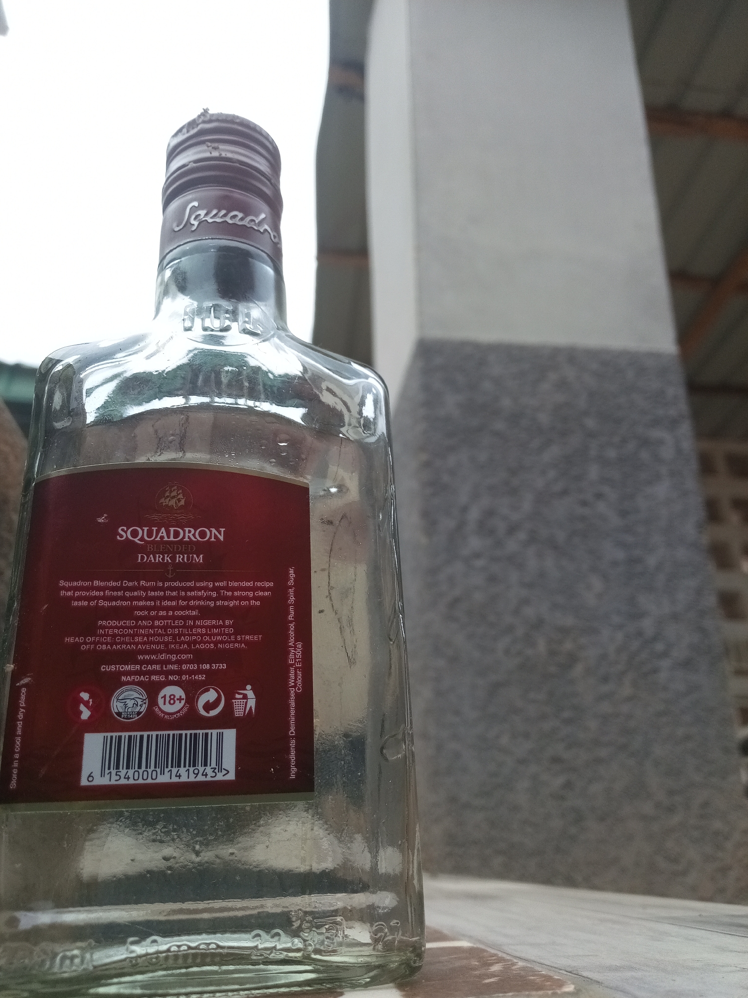 A close-up, eye-level shot shows the upper portion of a clear glass bottle of Squadron Blended Dark Rum, set against a blurred background. The bottle's neck is capped with a ridged, dark brown closure that has the word "Squadron" embossed in a script font. Below the neck, the clear glass transitions to a dark red label. The label features the brand name "SQUADRON" in large white lettering, followed by "BLENDED" and "DARK RUM" in smaller white text. A ship graphic is positioned above the brand name. Text on the label details the rum's production and composition: "Squadron Blended Dark Rum is produced using well blended recipe that provides finest quality taste that is satisfying. The strong clean taste of Squadron makes it ideal for drinking straight on the rock or as a cocktail. PRODUCED AND BOTTLED IN NIGERIA BY INTERCONTINENTAL DISTILLERS LIMITED HEAD OFFICE: CHELSEA HOUSE, LADIPO OLUWOLE STREET OFF OBA AKRAN AVENUE, IKEJA, LAGOS, NIGERIA. WWW.lding.com CUSTOMER CARE LINE: 0703 108 3733 NAFDAC REG. NO: 01-1452 Ingredients: Demineralised Water, Ethyl Alcohol, Rum Spirit, Sugar, Colour: E150(a)". Below this text, there are several icons: a red circular symbol with a ship inside a cogwheel, an "18+" symbol indicating an age restriction, a recycling symbol, and a waste bin symbol. A barcode is also present. On the far left of the label, text in a vertical orientation reads: "Store in a cool and dry place". The bottle is positioned on a surface that appears to be a tiled or bricked table. The background is out of focus, showing a large, light-colored pillar on the right, and hints of a ceiling structure and possibly vegetation on the left. The lighting is natural and appears to be daylight. The setting is outdoors, possibly in a yard or patio area, in New Stadium, Nigeria. There are no people or animals visible in the image.
