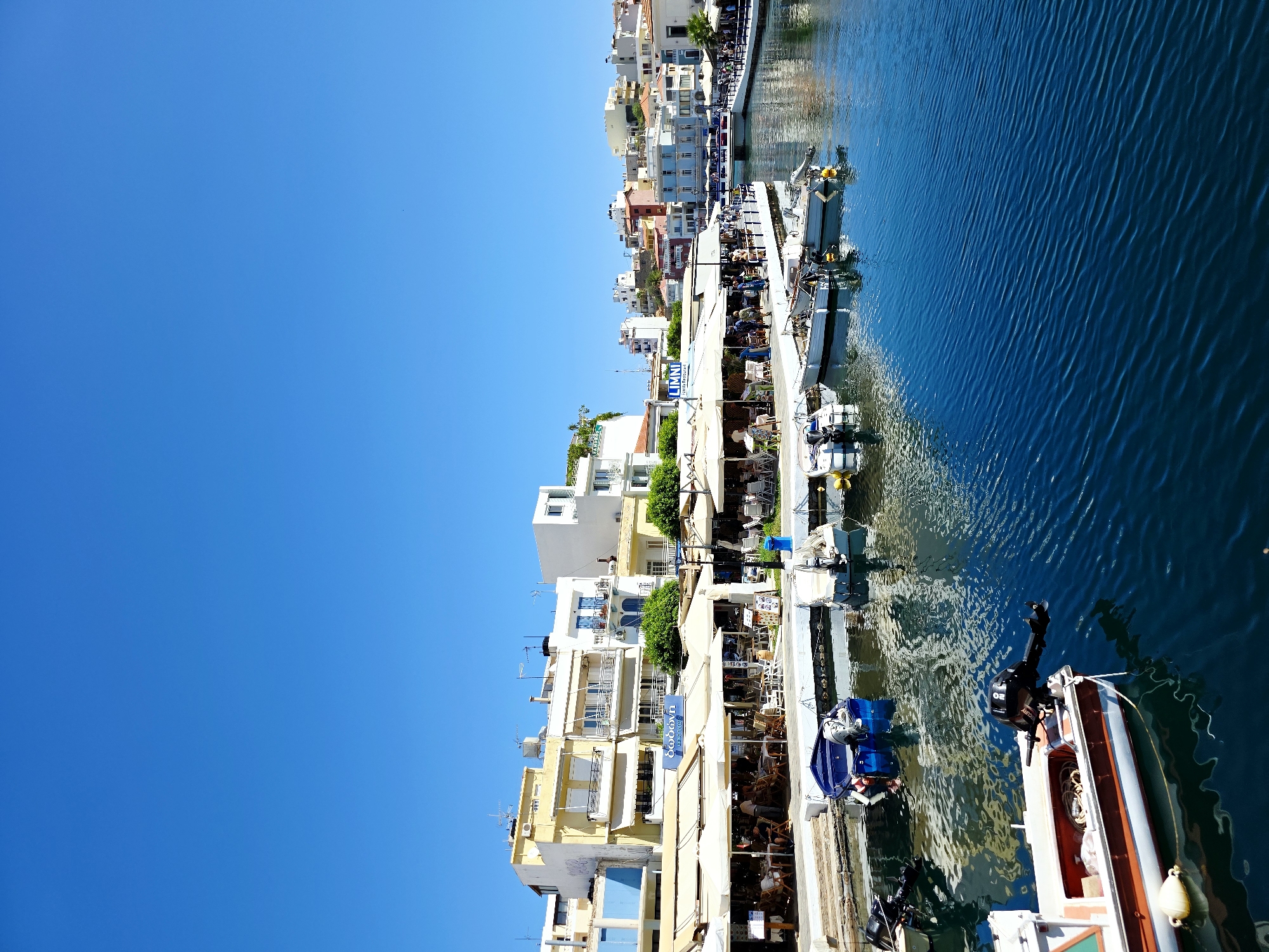 The image depicts a waterfront scene in Agios Nikolaos, Greece. Several boats are moored along a canal, with a row of buildings lining the opposite bank. These buildings appear to house restaurants or cafes with outdoor seating areas, many covered by awnings. People are visible seated at tables under the awnings. The water is a deep blue, with reflections of the buildings and sky visible on its surface. The sky is clear and bright blue, suggesting a sunny day. The architecture of the buildings suggests a Mediterranean or Greek style, with a mix of colors and balconies. The overall impression is one of a lively and picturesque waterside town.