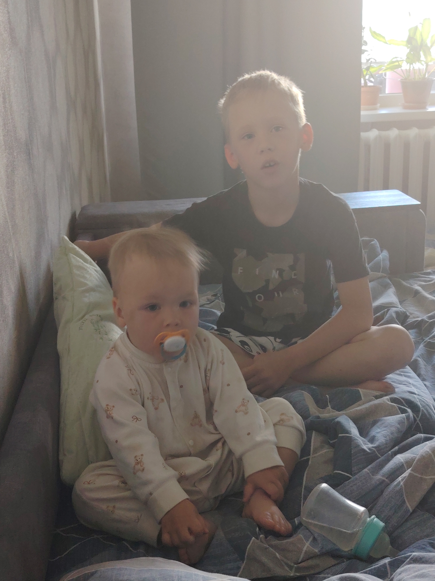 Two young children, possibly brothers, are sitting on a bed covered with a blue and gray patterned comforter. The younger child, wearing a white onesie with a teddy bear pattern and a pacifier, sits in front. The older child, wearing a dark T-shirt and patterned shorts, sits behind and to the right. A baby bottle lies on the bed near the younger child. The bed is positioned next to a wall with textured wallpaper. A pillow leans against the wall. A window with houseplants is visible in the background. The location is Chaikovsky, Russia.