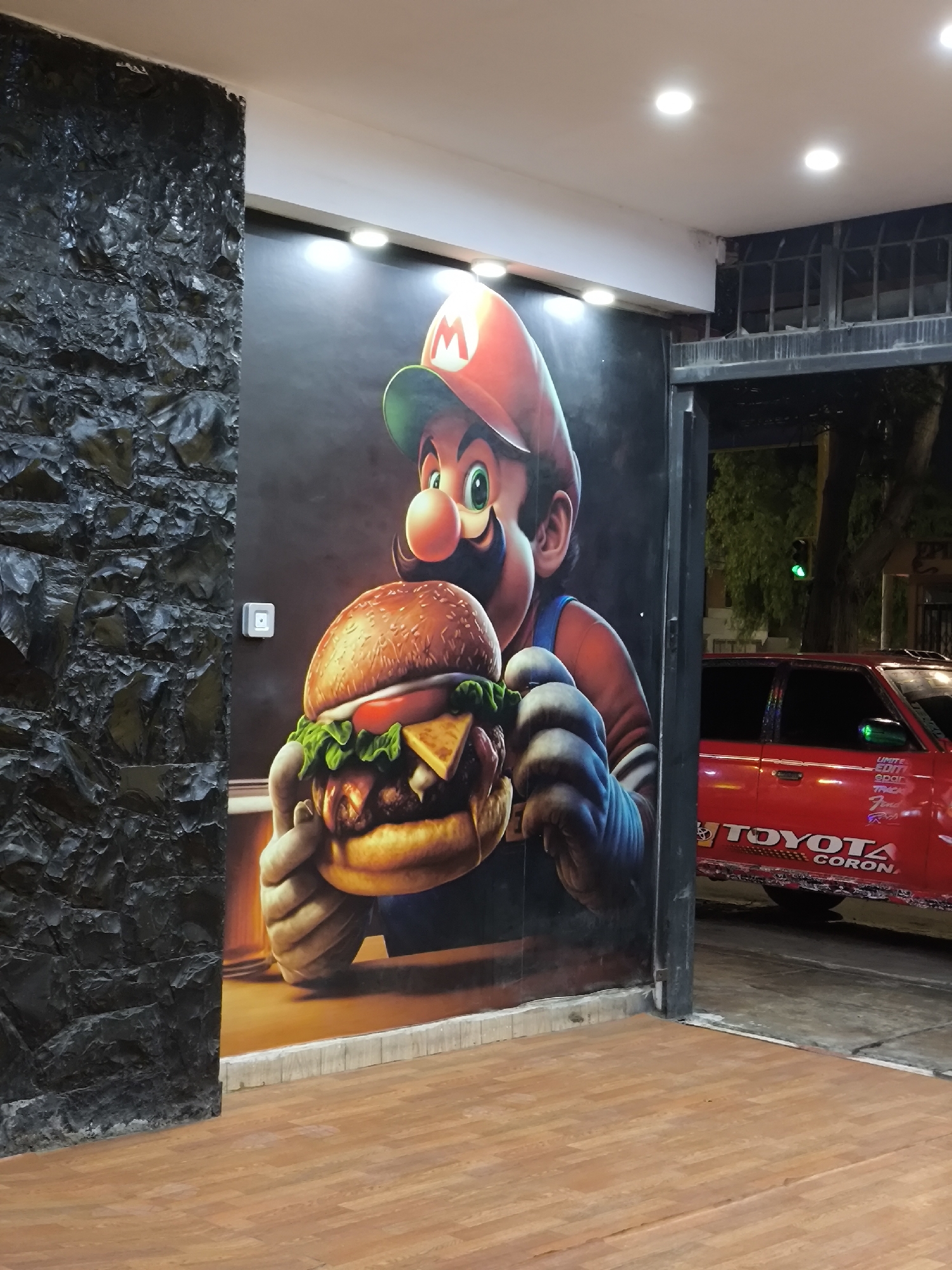 The image shows the exterior of a building, presumably in the city of Unknown, Unknown. A wall mural of Mario holding a hamburger is prominently displayed on a dark background. Adjacent to the mural is a textured wall with a rocky appearance. The flooring in front of the mural is composed of wood-like planks. Through an opening, a red Toyota car is partially visible, with writing on the side and the words "TOYOTA CORONA" identifiable. A green traffic light is discernible in the background.