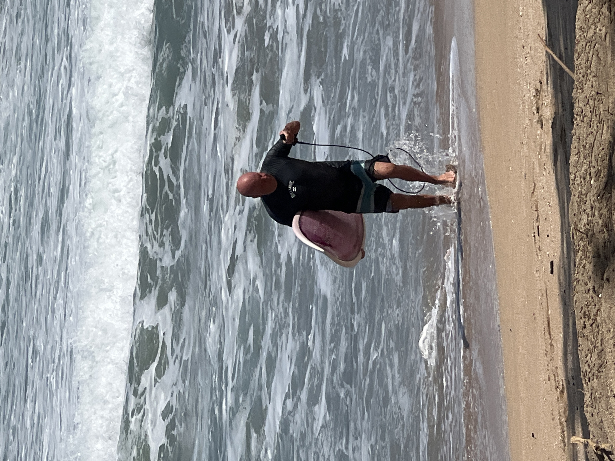 A bald man wearing a black rash guard and striped shorts is walking out of the ocean in Kuta, Indonesia, holding a pink and white boogie board. He is also holding a black leash connected to the boogie board. The water is choppy with white waves crashing behind him. The beach in the foreground is sandy with some driftwood.