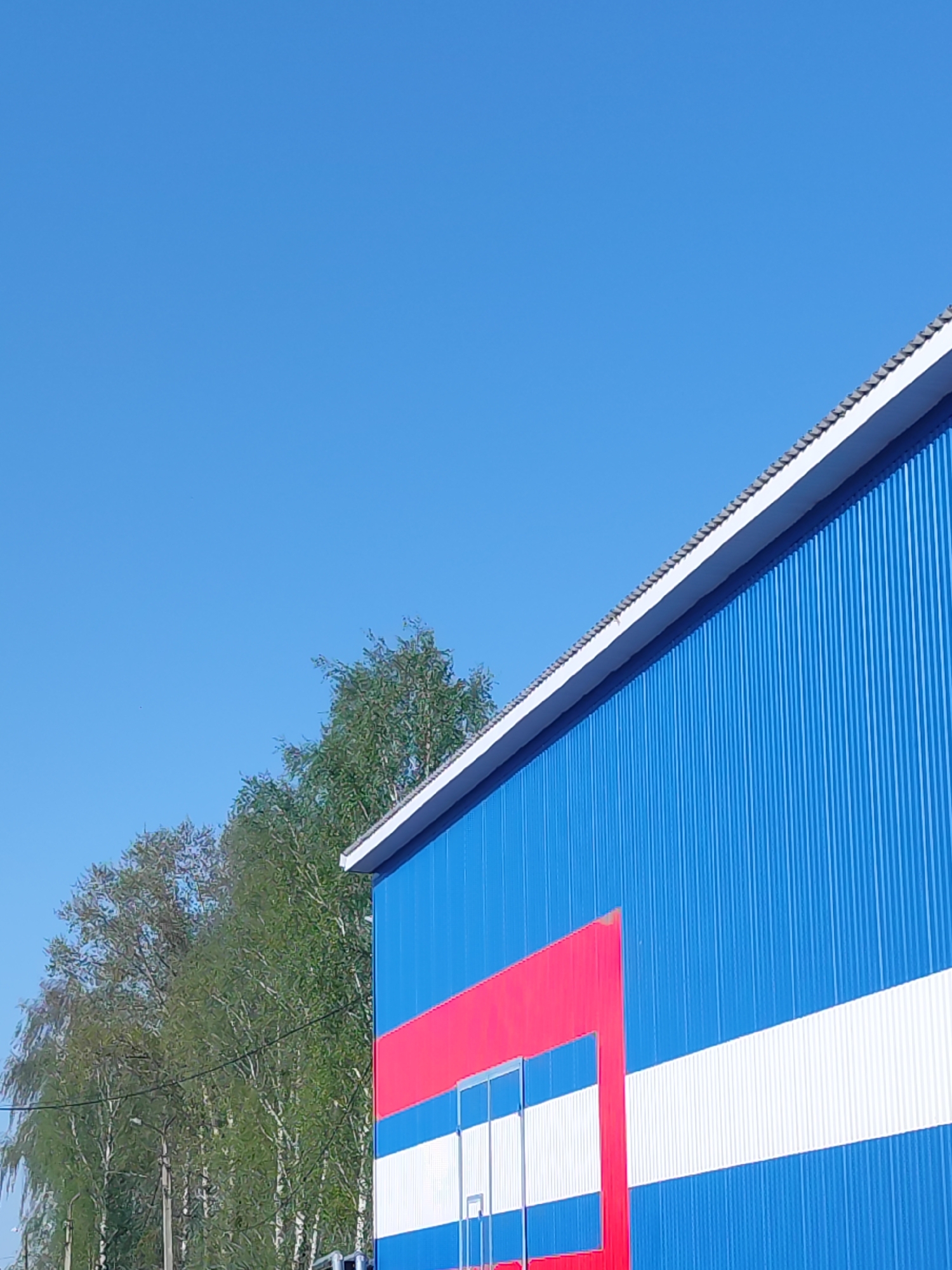 The image shows the side of a building under a clear, blue sky. The building's exterior is primarily blue with vertical ribbed metal siding, accented by a horizontal white stripe. A red section frames what appears to be a doorway, with a portion of the doorway also painted white. A row of trees with green foliage is visible along the left side of the frame, behind the building. The location appears to be in a suburban or industrial area.