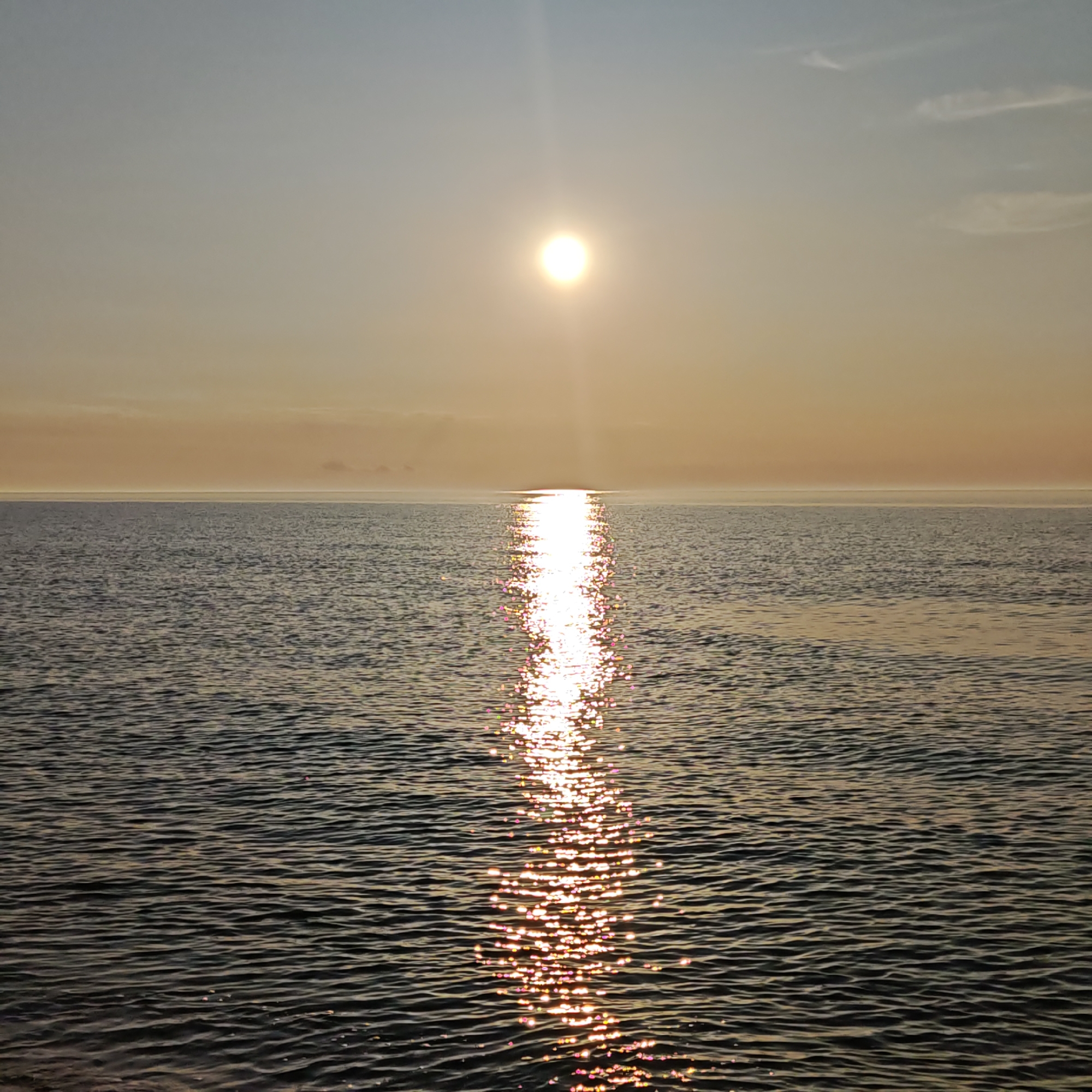 The image depicts a body of water under a bright, yellow sun. The sun is positioned high in the sky, casting a vertical line of shimmering light across the water's surface. The water itself appears slightly choppy, with small waves creating a rippled texture. The sky is a gradient of light blues and oranges, indicating either sunrise or sunset. There are a few faint clouds visible. The location in the city is Unknown, Unknown.