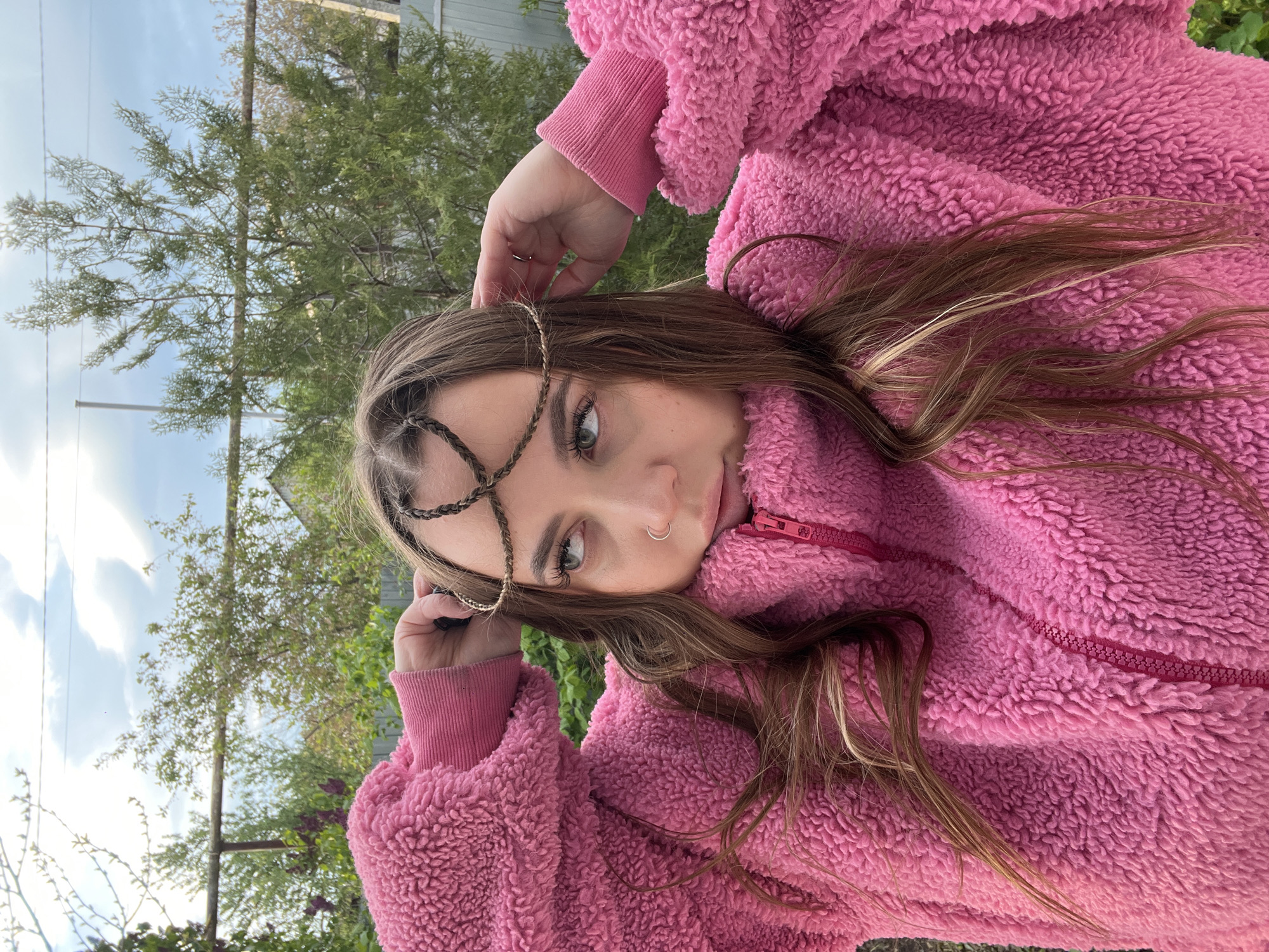 A young woman is pictured outdoors. She is wearing a fuzzy pink jacket and has a golden hair accessory consisting of braids crossing over her forehead. She is looking directly at the camera with a slight tilt of her head. Her hand is raised near the hair accessory. The background includes trees and a glimpse of the sky. The location is Novopokrovskaya, Russia.