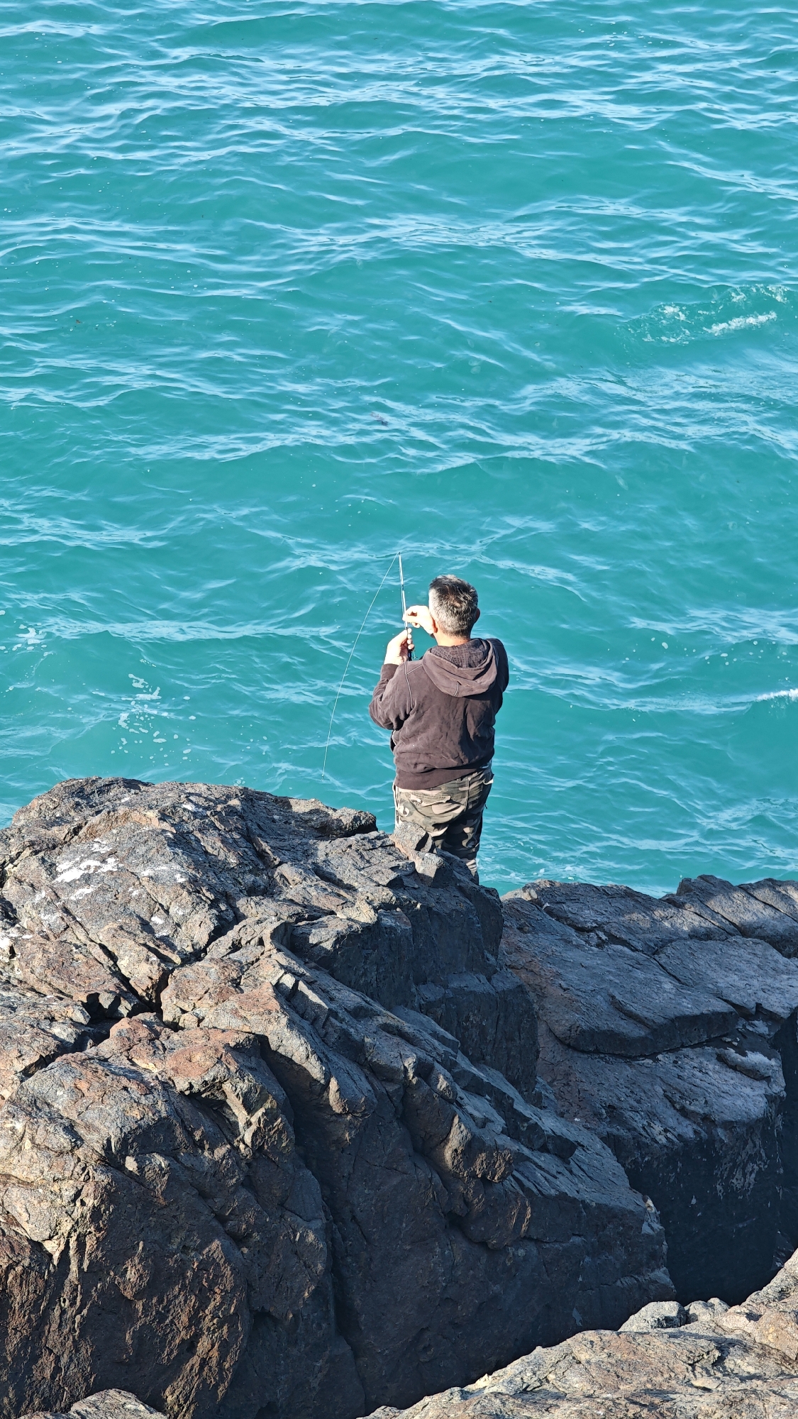 A man stands on a rocky outcropping, fishing in the sea near Sozopol, Bulgaria. The man faces away from the viewer, dressed in a dark hooded sweatshirt and camouflage pants. He holds a fishing rod, with the line extending into the turquoise sea. The rough, dark rocks form a jagged shoreline. The sea is relatively calm, with small ripples on the surface.