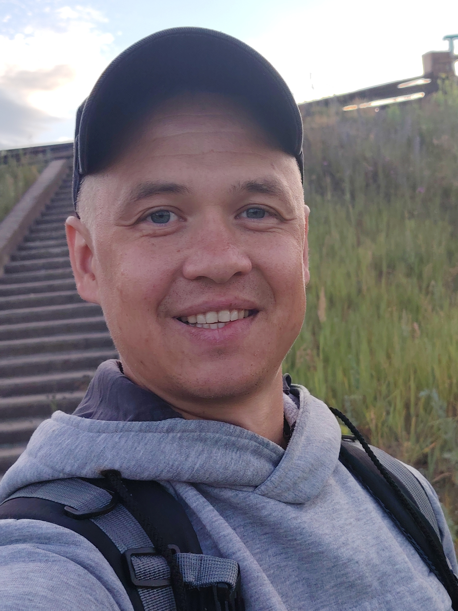 A man in a grey hooded sweatshirt and a black baseball cap is taking a selfie outdoors. He has fair skin, blue eyes, and a short haircut. He is smiling broadly, revealing his teeth. Behind him, a set of concrete stairs leads upwards, and a grassy embankment with some vegetation and what appears to be a railway or bridge structure is visible in the background. The sky is cloudy. The image appears to be taken at sunset. The location is the embankment near the Kama River in Chaikovsky, Russia, specifically on the staircase leading to the observation deck.