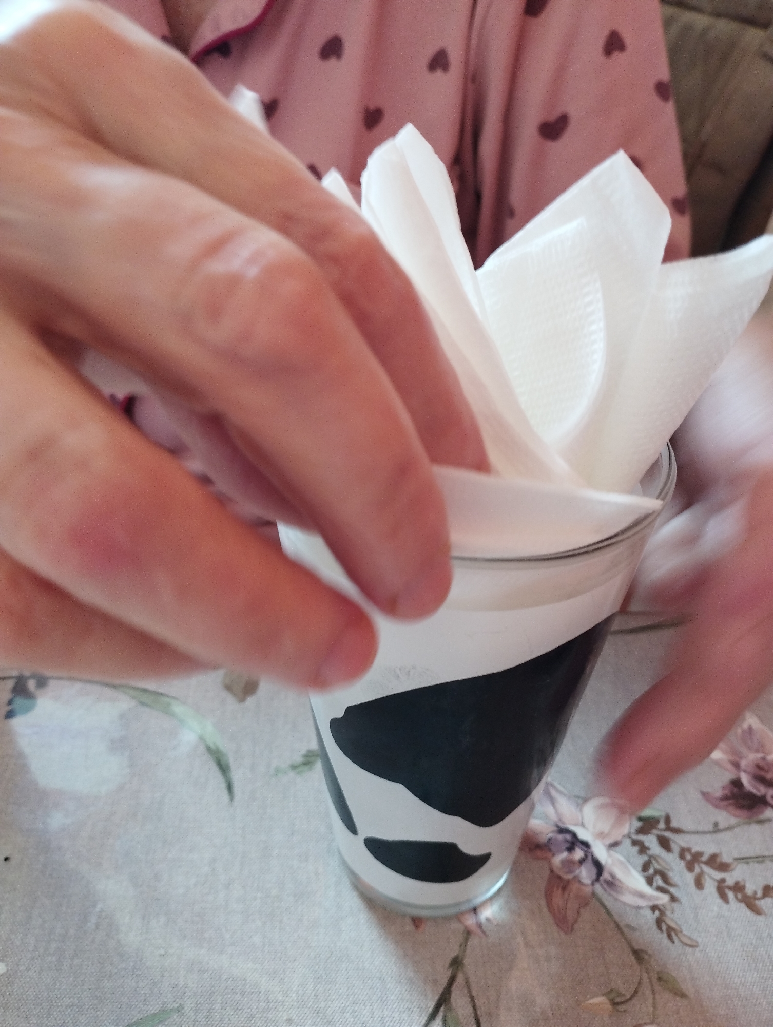 A close-up, indoor shot shows a person's hands interacting with white paper napkins arranged in a glass. The glass is decorated with a black and white cow-like pattern. The background consists of a pink garment with dark heart patterns and a tablecloth with floral designs. The scene appears to be a personal, domestic setting, possibly a kitchen or dining area. There is no information in the image to suggest a specific location in Moscow, Russia.