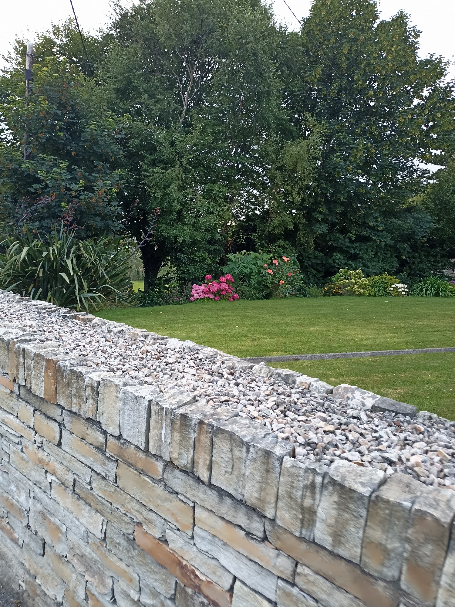 In Annagry, Ireland, a stone wall with a gravel top runs along the foreground, its rough-hewn stones displaying natural variations in color from beige to grey. Behind the wall, a manicured green lawn stretches towards a backdrop of lush trees. Clusters of vibrant pink and orange flowers are visible amidst the greenery, adding splashes of color to the serene landscape. The scene is bathed in natural light, suggesting an outdoor setting.