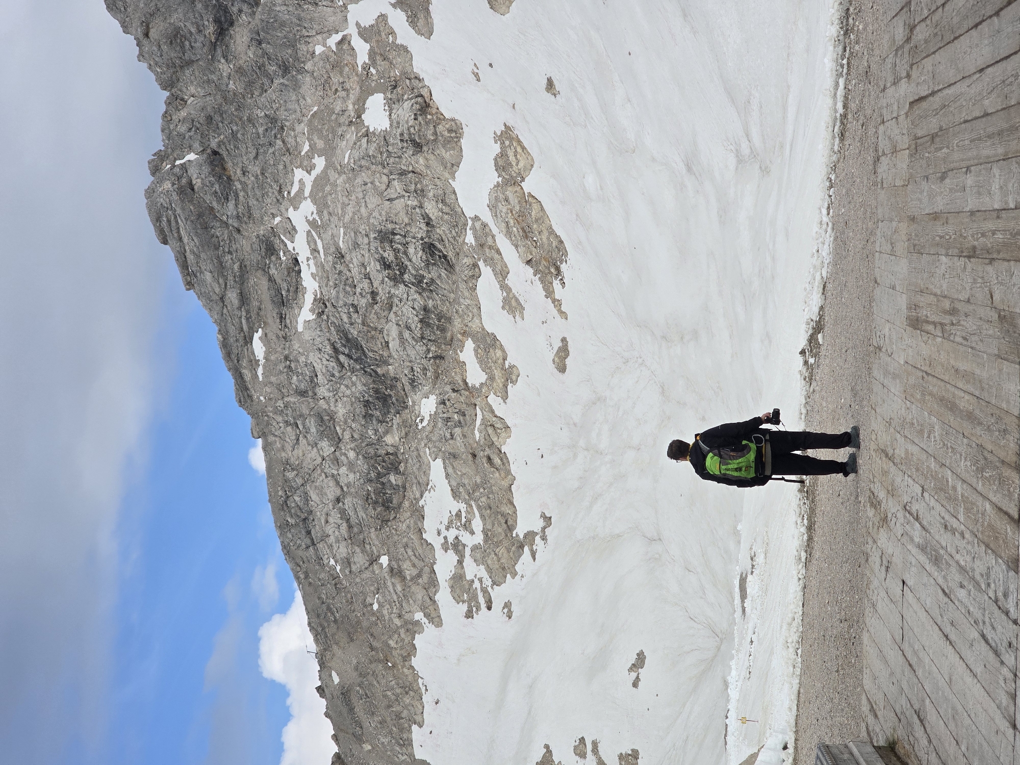 A person stands on a wooden platform facing a vast snowy mountain slope under a cloudy sky. The person is wearing a black jacket, black pants, and a bright green backpack. They are holding a camera in their right hand. The mountain is rocky and steep, with patches of snow scattered across its surface. The wooden platform appears to be part of a viewing area or trail. The scene is set in a high-altitude mountainous environment, likely the Zugspitze area near Garmisch-Partenkirchen, Germany, given the characteristic rocky peaks and snowfields. The lighting suggests it is daytime, possibly overcast.