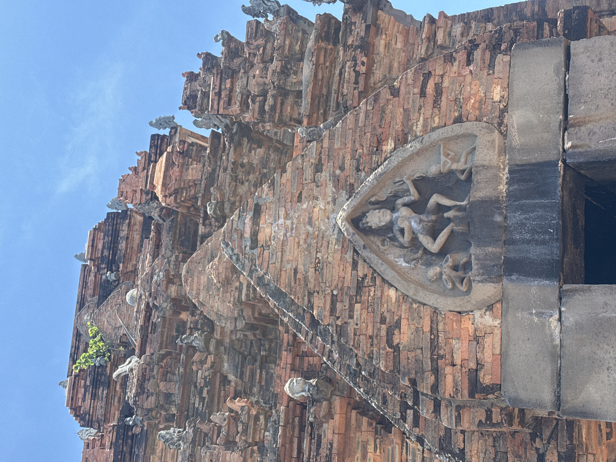 This is a close-up, upward-facing photograph of the Po Nagar Cham Towers in Nha Trang, Vietnam. The ancient brick structure dominates the frame, with the bright blue sky visible in the upper portion. The towers are adorned with intricate carvings and sculptures, some of which are weathered and partially damaged. In the center of the image, a triangular relief depicts a multi-armed deity with smaller figures on either side. Numerous smaller decorative sculptures of human-like figures and mythical creatures are scattered across the tiered brickwork. A few small plants are growing out of crevices in the brick. The overall impression is one of historical grandeur and natural decay.