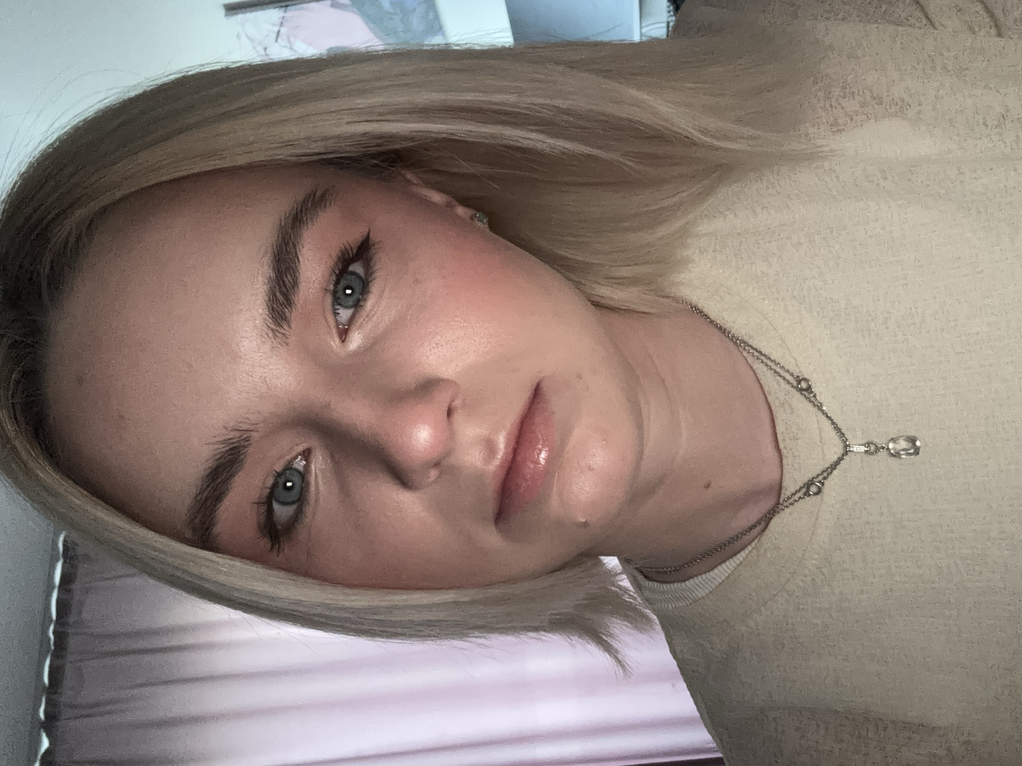 A close-up portrait shows a fair-skinned person with blonde, shoulder-length hair and light blue eyes. The person has well-defined eyebrows, a light application of makeup emphasizing their eyes, and glossy lips. They wear a light-colored knitted sweater and a delicate silver necklace with a small crystal pendant. The background is indistinct, showing a hint of a pink curtain or sheet and a pale wall. The photo is rotated, with the person's face tilted to the left. The image does not contain any indication of being taken in Ladozhskoe Ozero, Russia.