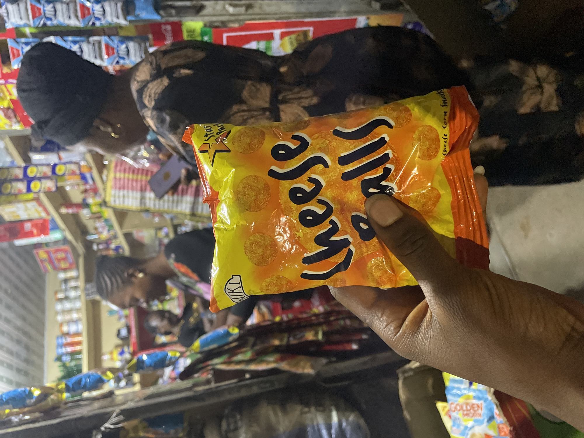 In a dimly lit convenience store in Jos, Nigeria, a person's hand holds a brightly colored bag of "Cheese Balls" snacks up to the camera. The bag, predominantly orange and yellow, features large, stylized text that reads "Cheese Balls," with smaller text below indicating "Cheese Corn Snacks." Behind the hand, the store's interior is visible, with shelves stocked with various food items and packaged goods. Two women are present in the background. One, wearing a black headwrap and a patterned dress, is looking down at her phone. The other woman, with braided hair and a colorful top, is standing near the shelves, also seemingly engaged with items in the store. The overall impression is a snapshot of everyday life within a small Nigerian business.