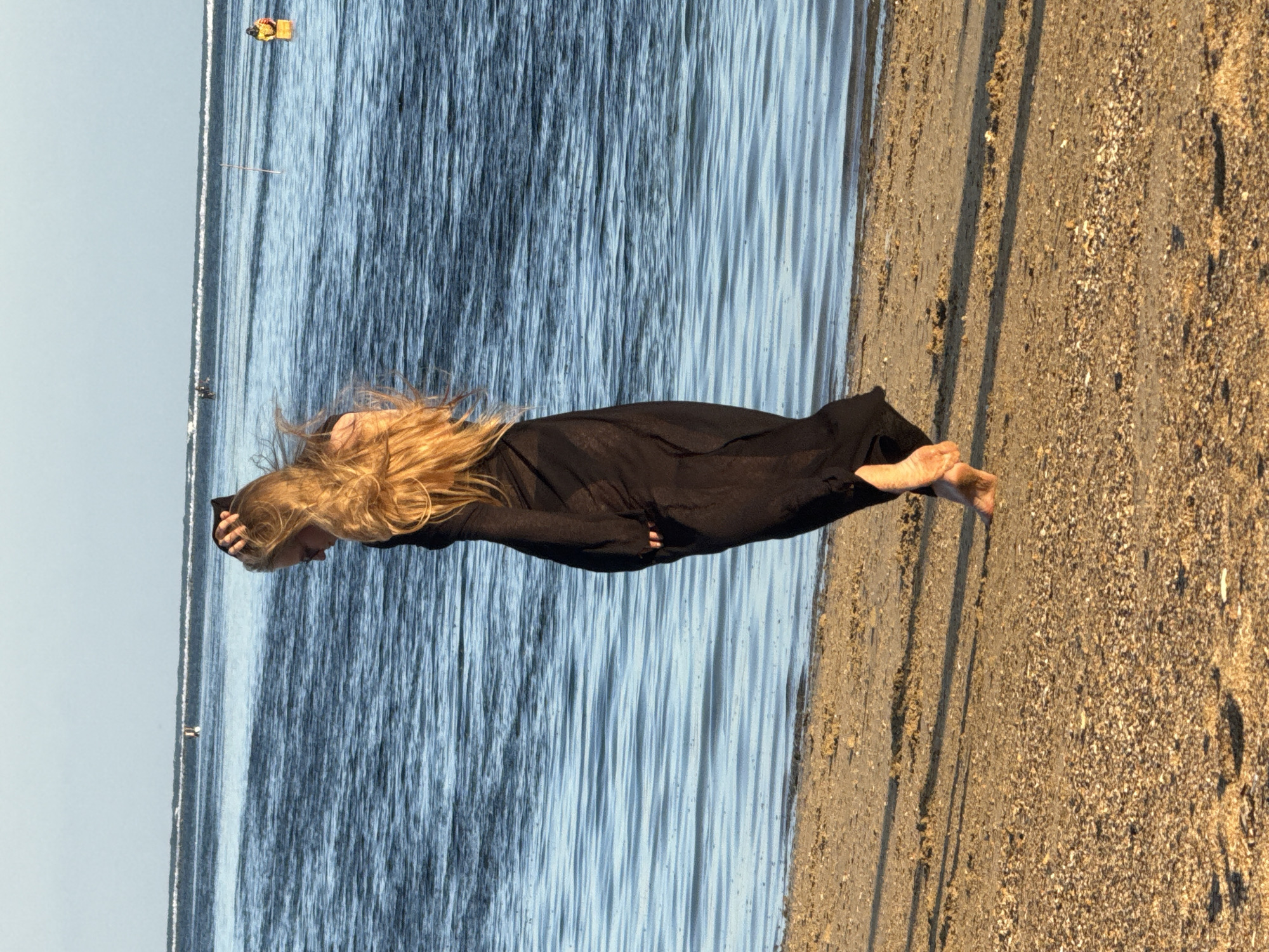 A young woman with long blonde hair, wearing a flowing black dress, walks along a sandy beach next to the ocean. The water is blue with gentle waves, and the sky is clear and light blue. The beach is covered in sand and small shells. In the distance, there are buoys and a few people in the water. The image appears to be taken in Denpasar, Indonesia.