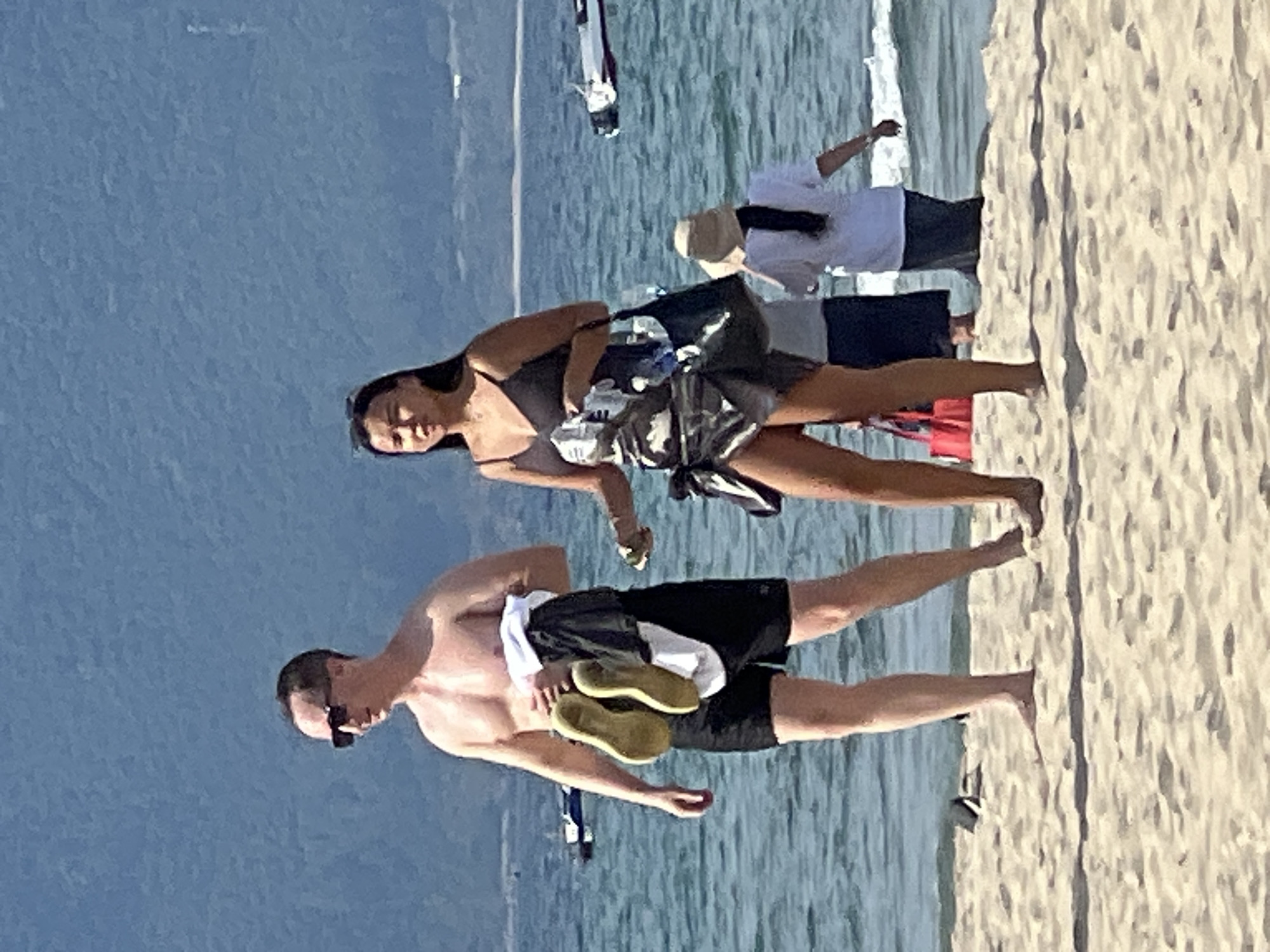 In Đà Nẵng, Vietnam, a man and a woman stand on a sandy beach near the ocean. The man, wearing black swim trunks and sunglasses, holds a white shirt and a pair of yellow sandals in his left hand. The woman, wearing a dark-colored swimsuit with a patterned sarong, has her left hand extended, holding a small object. Behind them, the turquoise ocean is visible, with several boats in the distance. Further back, a hazy blue mountain range forms the backdrop. Two other individuals are partially visible further down the beach, one wearing a white t-shirt and a hat, and another in dark clothing. The sand is light brown and appears undisturbed.