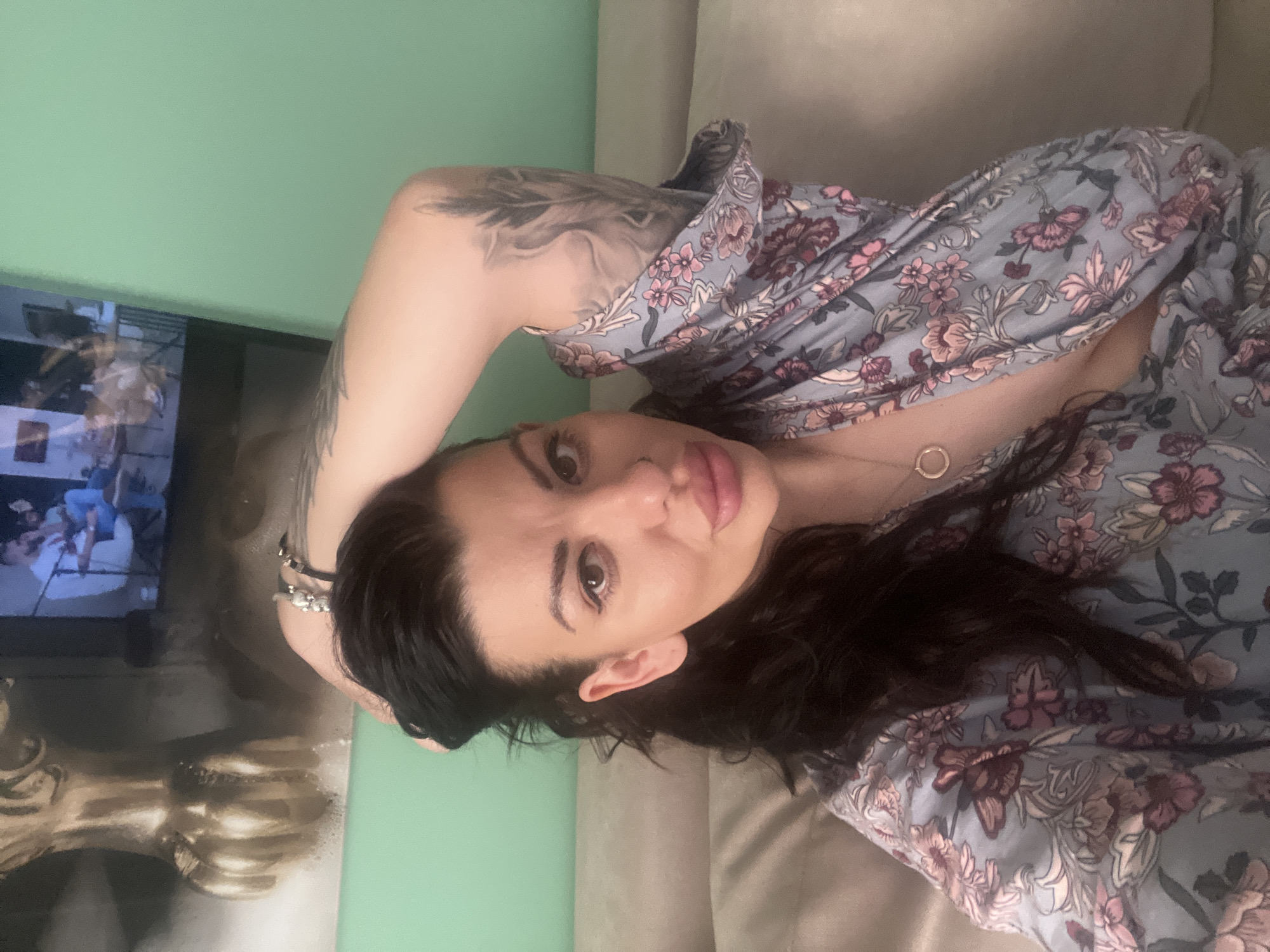A woman is lying on a couch in an indoor setting. She has dark hair and is wearing a floral-patterned shirt. Her left arm is raised, revealing a tattoo on her upper arm. She is looking at the camera. In the background, a television displays an image, and there is a painting or artwork on the wall. The room has a green wall. The location is not discernible as Plovdiv, Bulgaria based on the visual information provided.