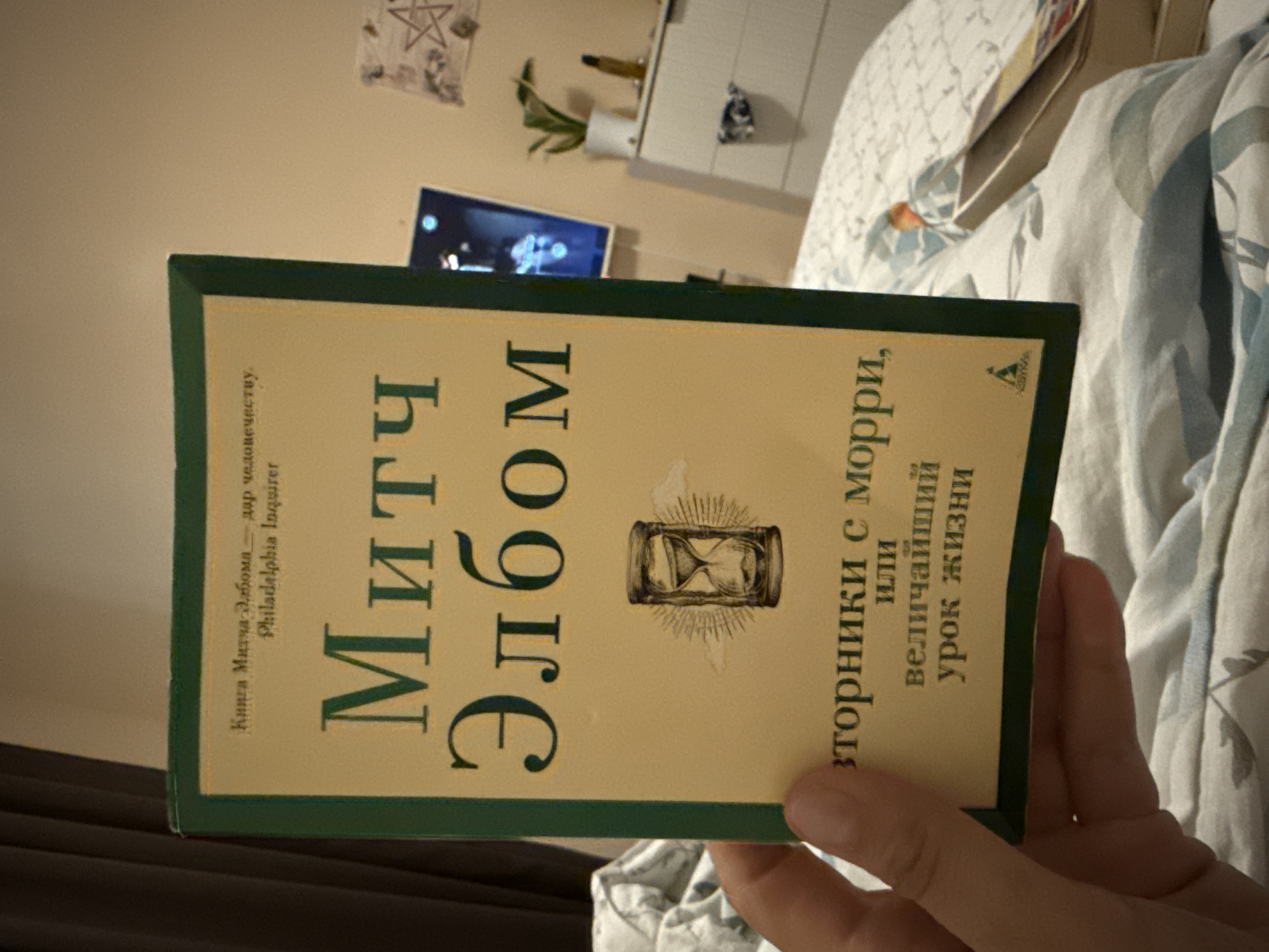 The image shows a person holding a book titled "Tuesdays with Morrie" (translated to Russian). The book cover is green with beige text, featuring the author's name "Mitch Albom" in large, stylized Cyrillic letters, an image of an hourglass, and the book title in smaller Cyrillic letters. The book also mentions that it is a Philadelphia Inquirer bestseller. The person's hand is visible in the foreground, holding the book against a blurred background of bedding and furniture. The background includes a television, dresser, and wall. There's no information to indicate it is located in Tobolsk, Russia.