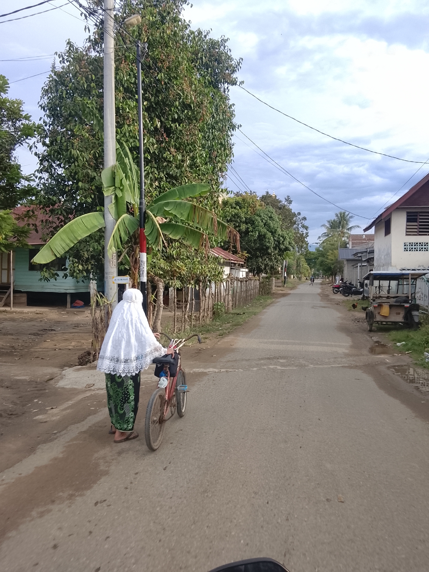 An eye-level, medium shot captures a rural street scene in Geunteng, Indonesia. A woman, partially obscured from the waist down by a bicycle, stands facing away from the camera, clad in a white hijab and a green and black patterned skirt. She is holding onto the handlebars of a red bicycle, which has a black bag attached to the rear rack. She is standing on a dirt shoulder next to a paved road. The road stretches into the distance, lined with wooden fences and various trees. Buildings, including a light blue house with a red roof and a white building with a red roof, are visible on either side of the road. A street light pole stands to the left of the woman, adorned with red and white signage. The sky above is overcast with clouds. Several motorcycles and a three-wheeled vehicle are parked along the right side of the road further down. A chicken is visible on the ground near the parked three-wheeled vehicle.