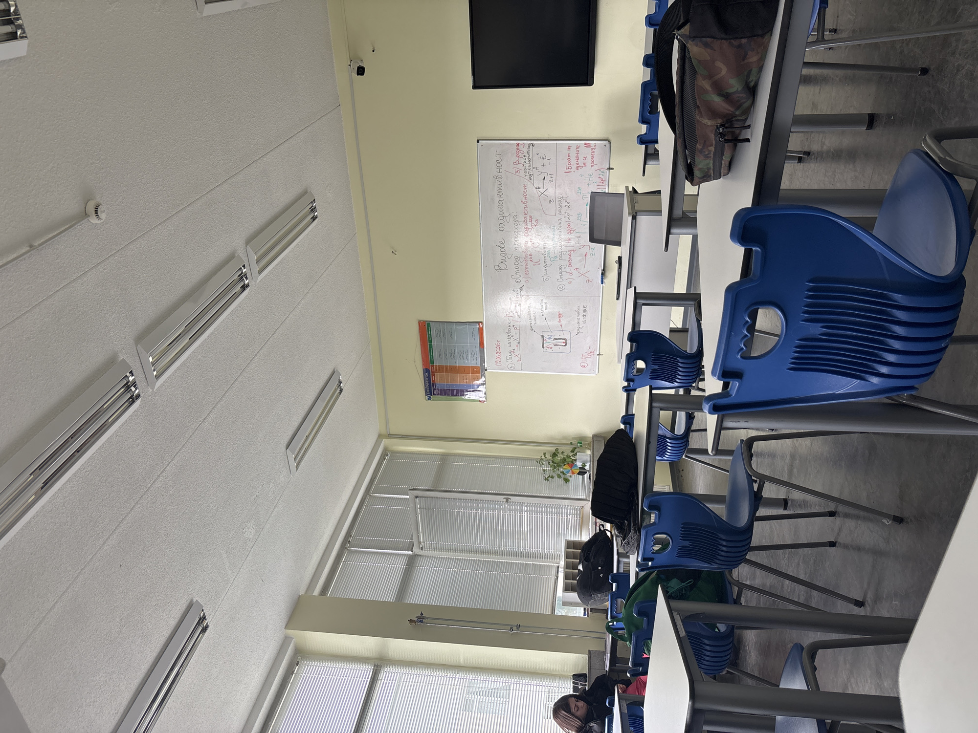 The image shows an indoor scene of a classroom or lecture hall, viewed from a slightly elevated perspective. The room is equipped with rows of white desks and blue plastic chairs. One person with dark hair is partially visible, seated at a desk in the lower right quadrant of the image.
The setting includes a white textured ceiling fitted with multiple long fluorescent light fixtures. A fire alarm or smoke detector and a small security camera are mounted on the ceiling and upper wall respectively. The walls are painted a light yellow. Large windows with horizontal blinds are visible along one side of the room.
A prominent whiteboard on the far wall displays text written in Bulgarian, titled "Видове радиоактивност" (Types of Radioactivity). The content includes subheadings such as "Според произхода" (According to origin) with "естествена радиоактивност" (natural radioactivity) and "изкуствена" (artificial), and "Според радиоактивния разпад" (According to radioactive decay) discussing "α-разпад" (alpha decay) and "Б-разпад" (beta decay), along with related nuclear equations and principles. A date, "02.06.2026г." (June 2nd, 2026), is also visible on the whiteboard. Above the whiteboard, a black flat-screen monitor is mounted. To the left of the whiteboard, a colorful wall chart or poster is displayed.
Other objects include backpacks resting on desks, such as a camouflage-patterned one in the mid-ground and a black one near the windows. A small potted plant and a decorative rainbow-colored item are placed on a sill or shelf by the windows. The room appears to be largely unoccupied, suggesting a quiet moment in an educational environment.