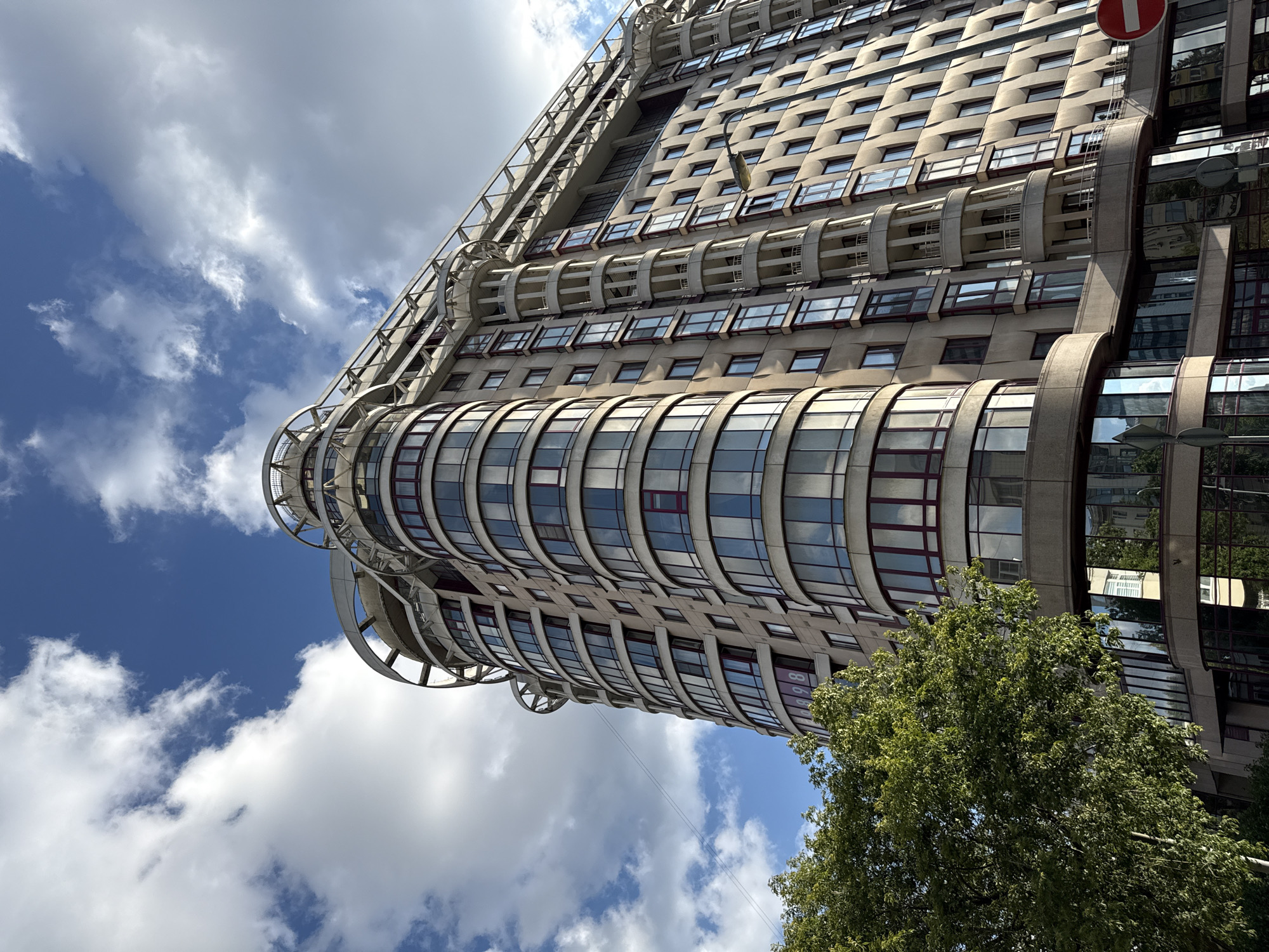 The image shows a modern, curved apartment building with a light-colored facade, located in Moscow, Russia. The building features numerous large, reflective windows with dark red frames and decorative balconies. The top of the building is adorned with circular architectural elements. A lush green tree is visible in the lower left portion of the frame, partially obscuring the building's base. The sky is blue with scattered white clouds. The scene appears to be captured during daylight hours.
