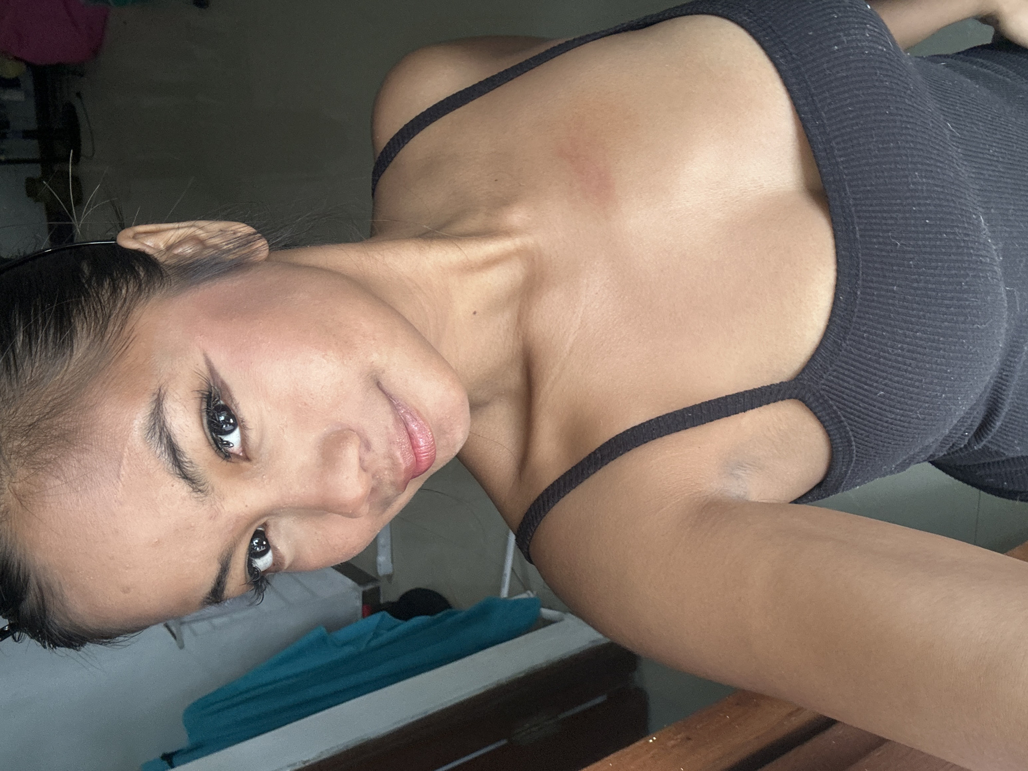 A young woman with dark hair and brown skin is taking a selfie indoors. She is wearing a black ribbed tank top. She has on makeup, including eyeliner and lip gloss. Her hair is pulled back, and a few strands are sticking out near her ear. There is a turquoise garment hanging on the left side of the frame, and a wooden chair or furniture is visible in the lower left corner. The background is dimly lit and appears to be a room in a house.