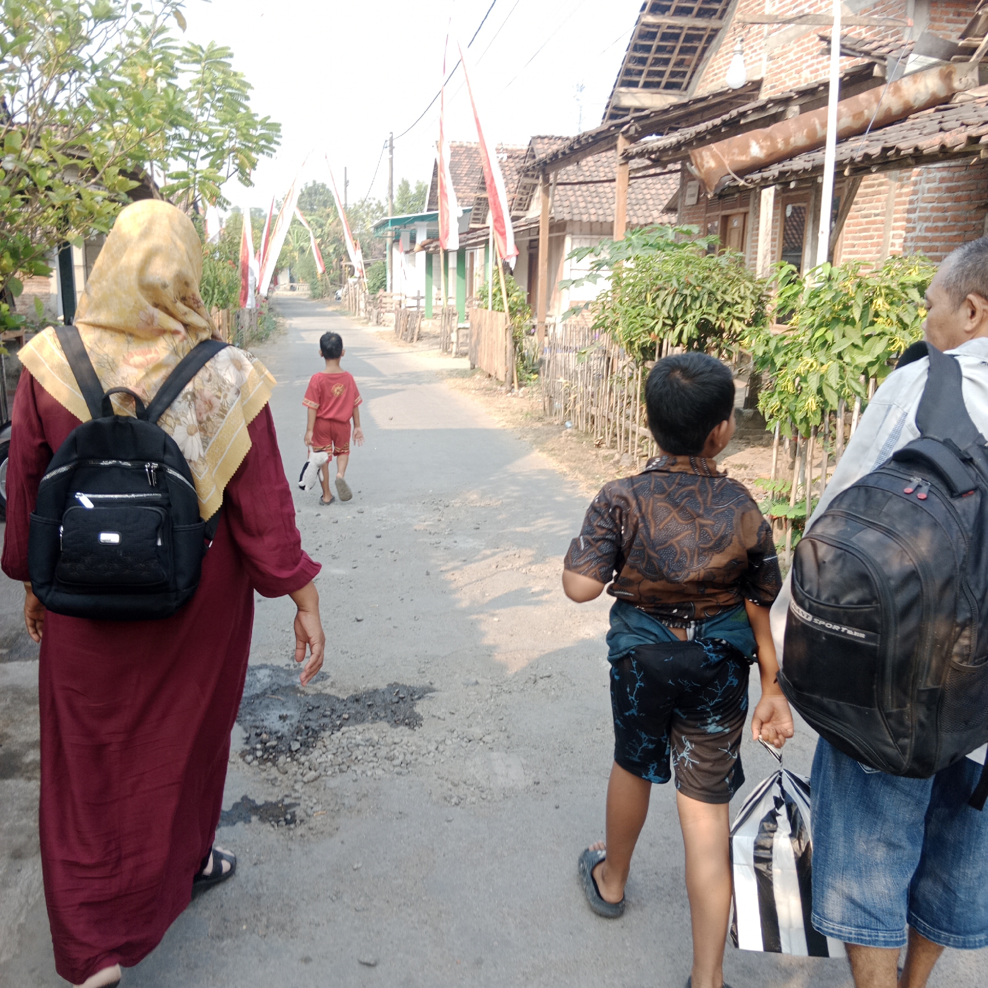 In the city of Ngringin, Indonesia, an overcast day frames a scene of three individuals walking along a narrow, dusty road lined with simple, traditional houses. A woman, her back to the camera, wears a maroon robe and a yellow headscarf, carrying a black backpack. Ahead of her, a young boy in a red shirt and shorts walks with a stuffed toy. To the right, a person with a grey shirt and blue denim shorts, also with a backpack, walks alongside the boy. Indonesian flags hang across the road, suggesting a local celebration or event. The houses exhibit weathered brickwork and corrugated iron roofs, with small gardens and wooden fences visible. The road itself is unpaved and appears to be a residential pathway.