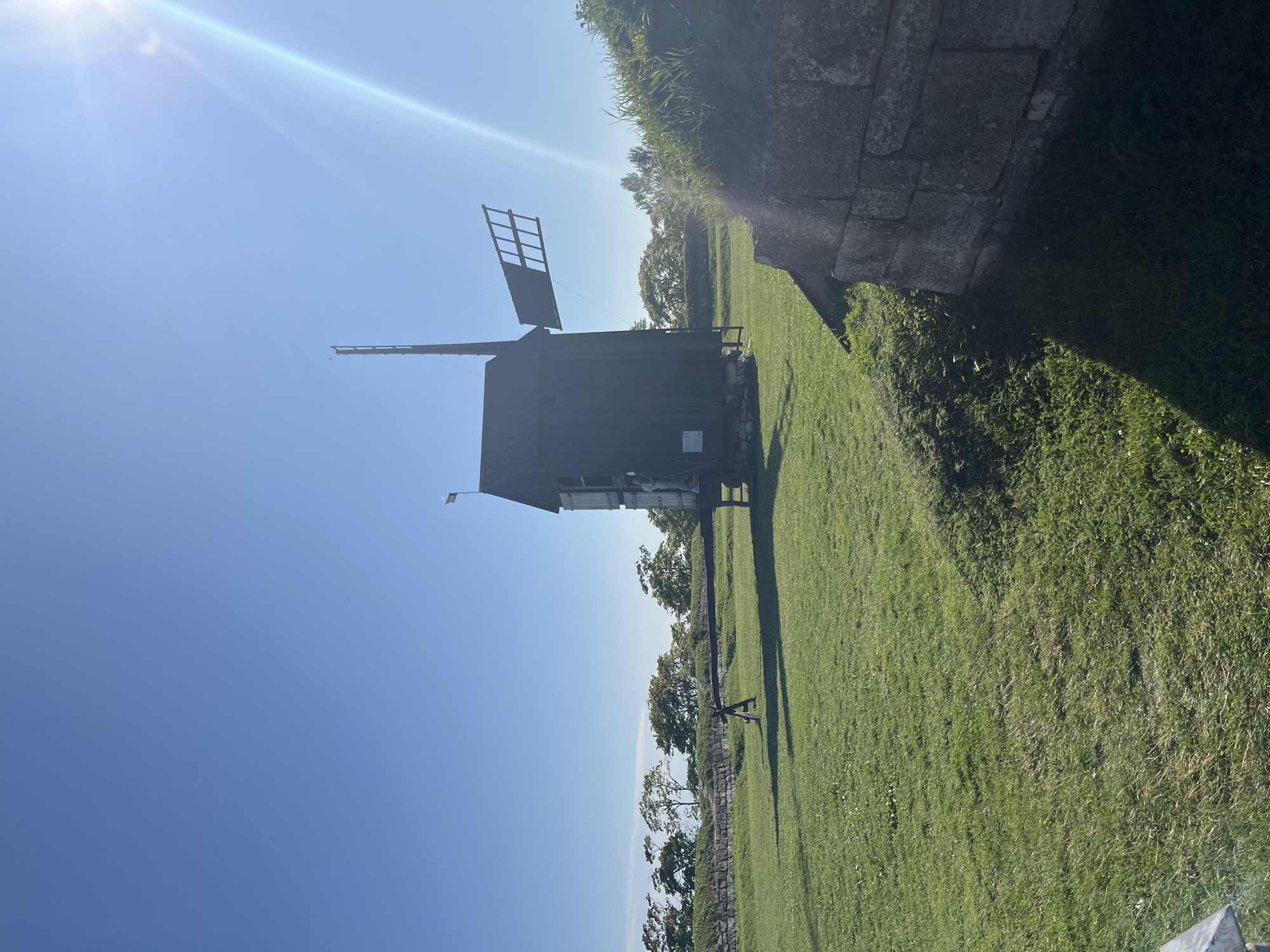 The image shows a dark wooden windmill with one visible sail, situated in a grassy field under a bright blue sky with a prominent sun flare. A stone wall is visible in the foreground and to the right. In the background, a line of trees and vegetation runs along a low, stone-capped embankment. Two people are visible near the windmill; one appears to be standing on a wooden platform attached to the windmill, and another is further away in the field, possibly operating or examining something on the ground. The scene is likely Kuressaare, Estonia, given the architectural style of the windmill and the surrounding landscape often associated with historical coastal fortifications.