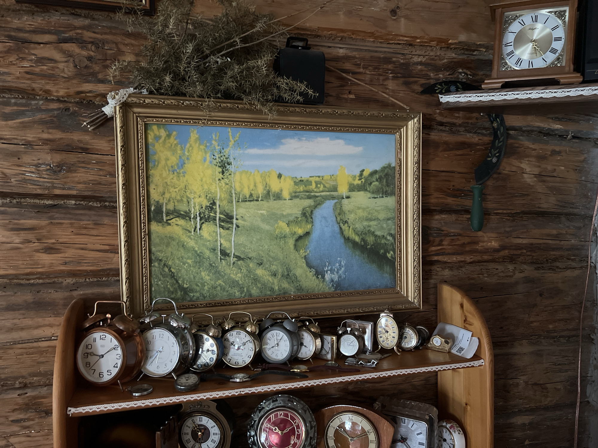The image depicts an interior scene within a rustic wooden building, likely a room in Norviliškės, Lithuania. The walls are constructed from rough-hewn logs.
The primary focus of the image is a collection of clocks displayed on wooden shelves. A large framed painting of a landscape with a river and trees hangs on the wall above the shelves.
On the top shelf, to the right of the painting, a square wooden desk clock with Roman numerals is visible. Below this, a decorative curved shelf holds several alarm clocks of various sizes and designs. These include large, metallic alarm clocks with twin bells on top, as well as smaller, ornate table clocks. A few pocket watches and what appears to be a small digital device are also present on this shelf.
The lower shelf displays more clocks, including several large, decorative mantel clocks with prominent Roman numerals and elaborate casings. One clock has a red face with gold accents.
Other items visible in the scene include a dark rectangular object, possibly a small case or speaker, placed on the wall above the painting, and a bundle of dried twigs tied with lace, also on the wall. A green-handled sickle with painted floral decoration hangs from the upper shelf. The overall impression is that of a collection of antique or vintage timepieces displayed in a traditional, rustic setting.