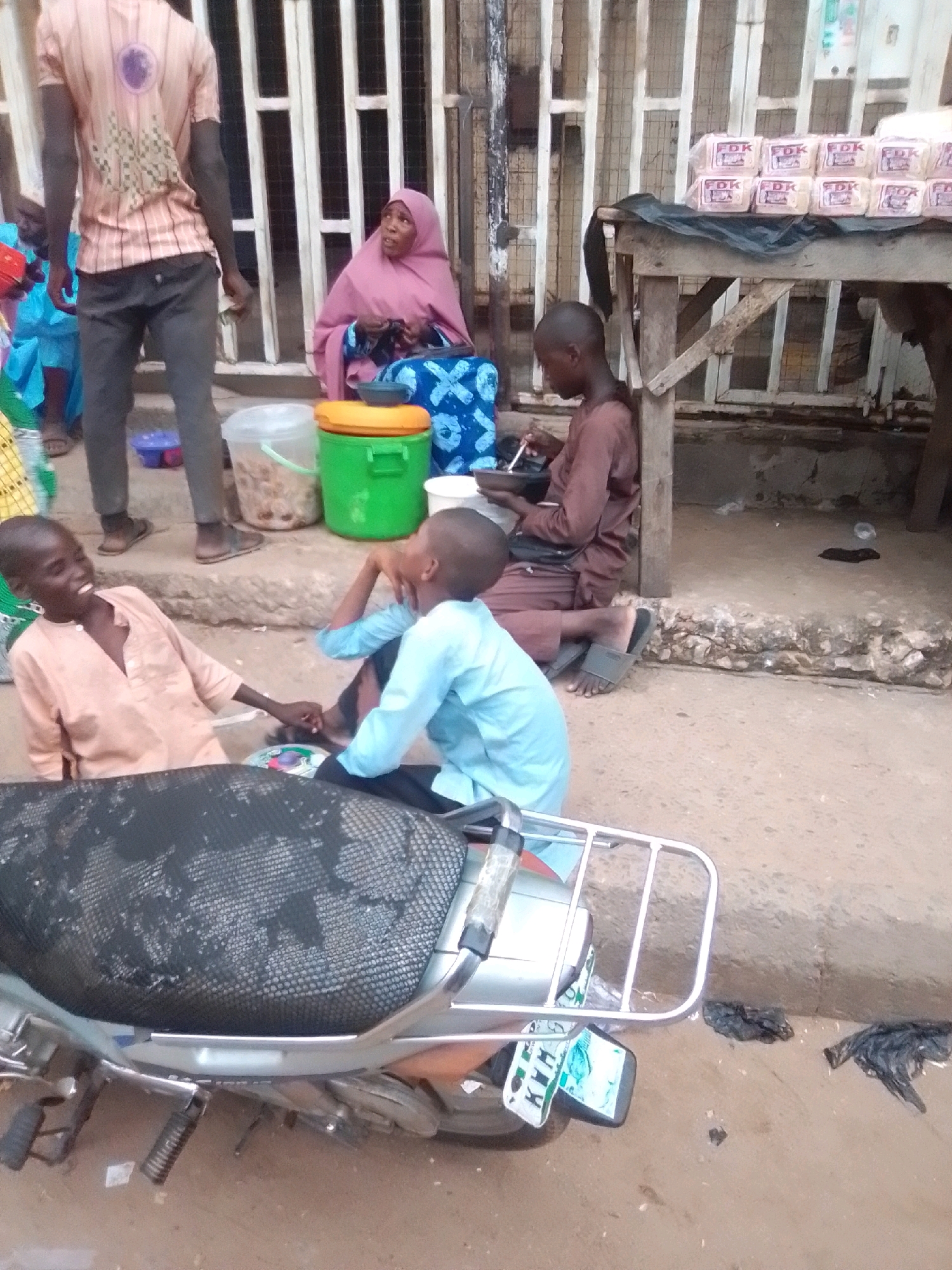 In Arawa, Nigeria, a street scene unfolds with several individuals and objects. A person wearing a pink and white striped shirt and dark pants stands near a vertical white gated structure. To their right, a woman in a pink hijab and patterned blue dress is seated. In the foreground, two young boys are interacting. One boy, wearing a peach-colored shirt, is seated and holding the hand of another boy in a light blue shirt, who is also seated. They appear to be engaged in a conversation or playful interaction. Beside them, a green cooler is visible. In the background, a table is laden with packages of bread. A motorcycle with a black seat and chrome accents is parked in the lower portion of the image, with its license plate visible. The ground is dusty, indicative of an outdoor urban environment. Debris, including black plastic bags, is scattered on the ground near the motorcycle.