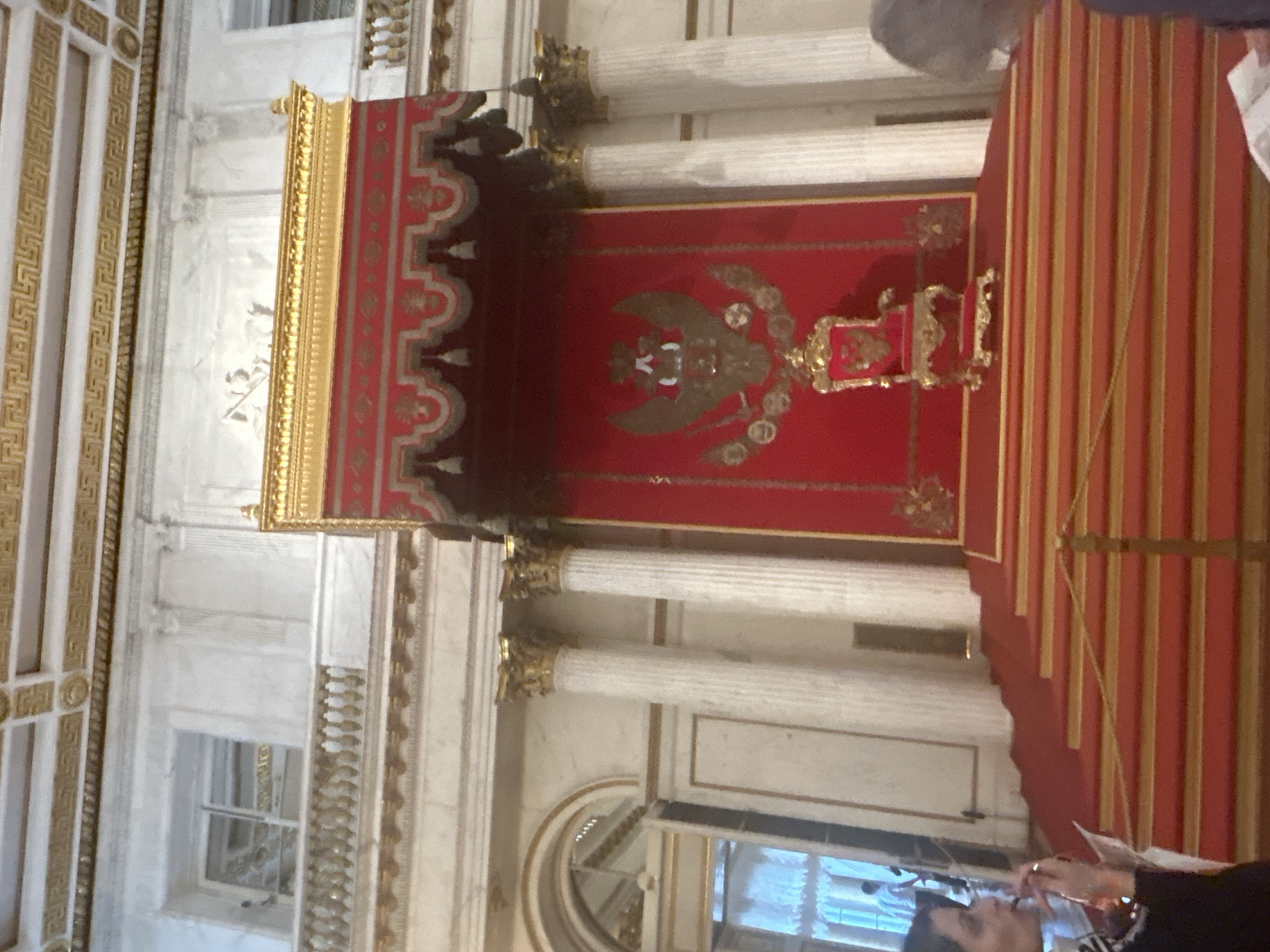 The image shows a grand throne room with a red throne placed on a raised platform with red carpeted steps. The throne is ornate and features a double-headed eagle crest. A red canopy with gold trim hangs above the throne. The room is decorated with marble columns and gold accents. Two people are partially visible in the foreground, one on the left looking to the right and holding a pamphlet, and another on the right also looking down at a pamphlet. The background shows architectural details of the room, including a balcony and a window. The scene appears to be a formal or historical setting.